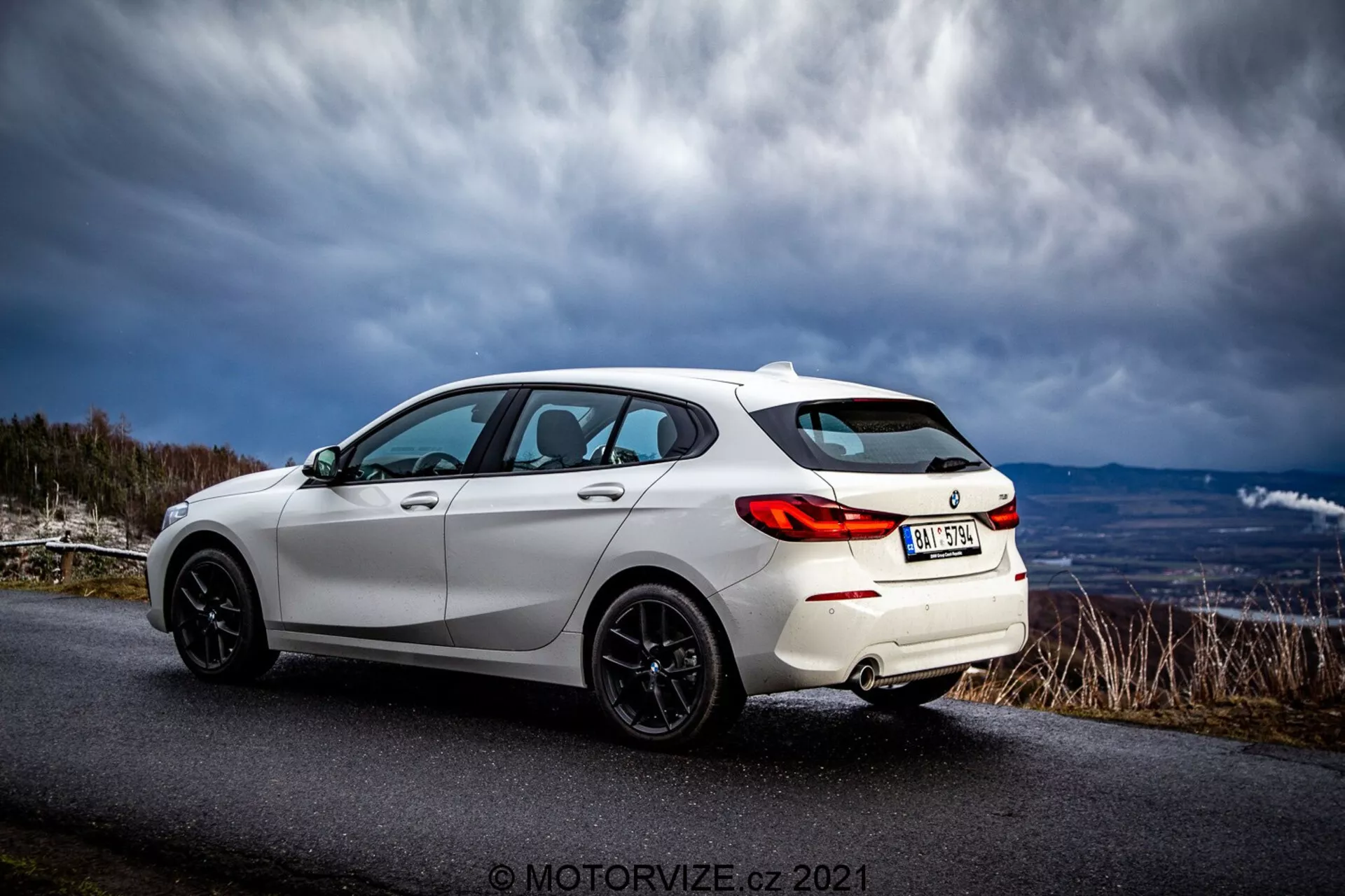 Un moderno hatchback blanco BMW 116i 2019 capturado desde una vista trasera de tres cuartos. Las características deportivas del exterior incluyen luces traseras LED, llantas de aleación negras de múltiples radios, un alerón trasero, ventanas tintadas y líneas de carrocería elegantes que resaltan su diseño aerodinámico. El vehículo está estacionado en una carretera asfaltada en una zona rural o montañosa bajo un cielo dramático.