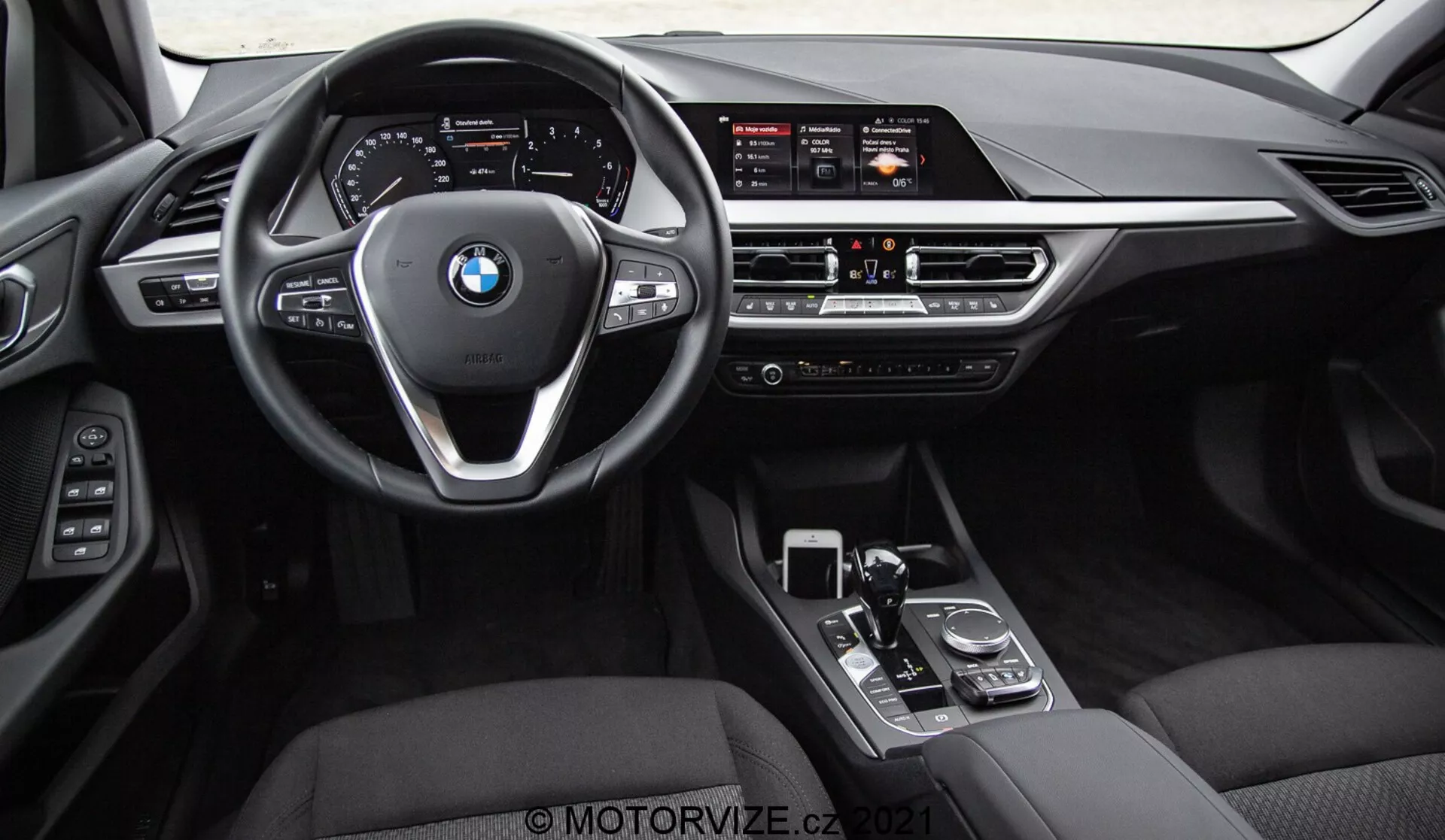 Vista interior de un BMW Serie 1 2019 (BMW 116i) tomada desde los asientos traseros, mostrando el tablero delantero completo, el volante con el logo de BMW y botones de control, y la consola central. Los elementos visibles incluyen la amplia pantalla de infoentretenimiento, controles manuales de clima, ventilaciones, palanca de cambios, dial giratorio y panel táctil. El interior cuenta con acabados en cuero negro o cuero sintético con elementos de acabado metálico y de alto brillo, enfatizando el lujo y la avanzada tecnología de interfaz del conductor.