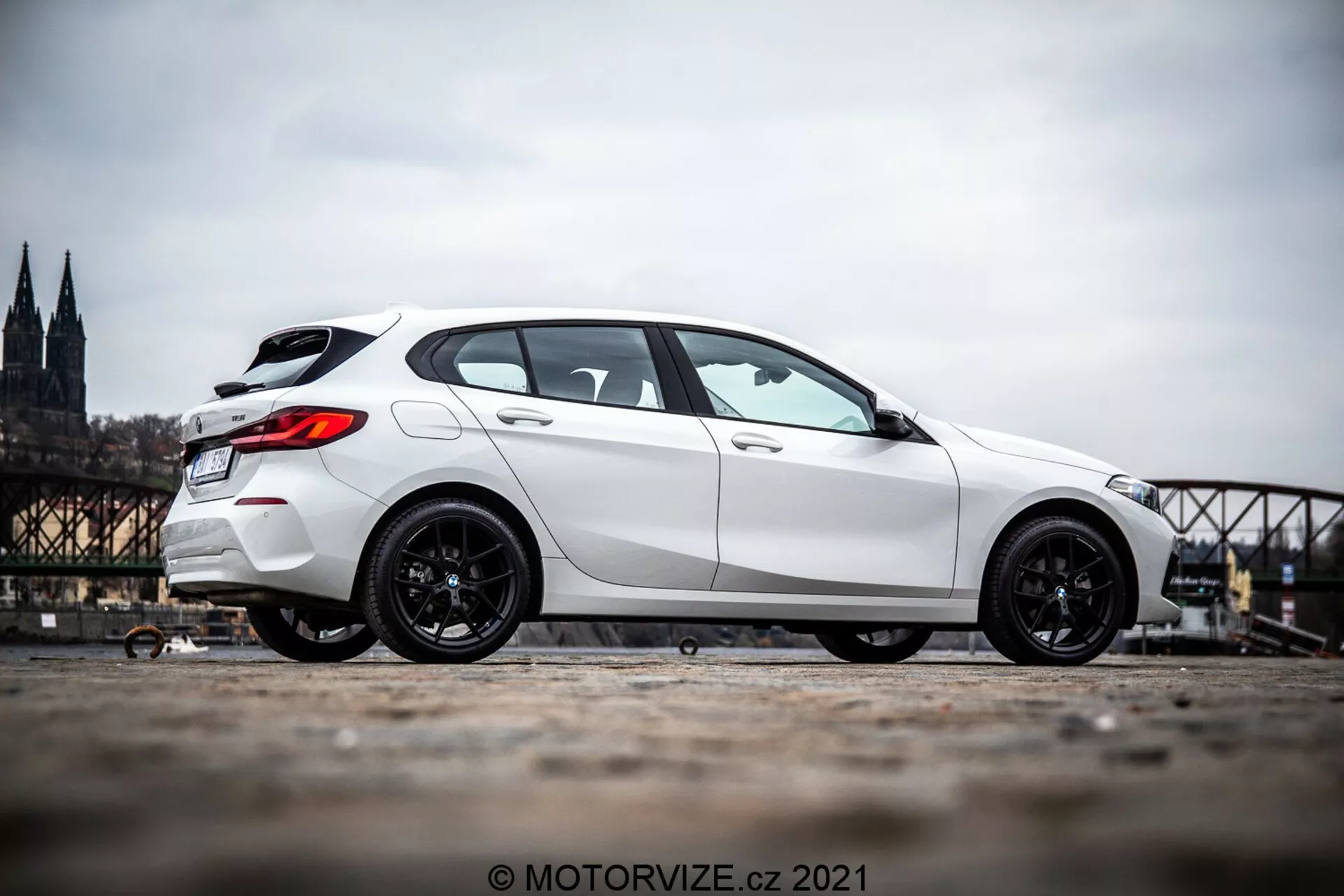 Un hatchback BMW Serie 1 (BMW 116i) blanco de 2019 fotografiado desde un ángulo lateral de perfil, mostrando el lado del vehículo desde justo delante de la rueda delantera hasta la parte trasera. El coche está posicionado en el lado izquierdo de la imagen con un fondo urbano histórico borroso que incluye una catedral y un puente. La imagen destaca las llantas deportivas de color oscuro del coche y su postura dominante debido a la perspectiva de nivel del suelo inferior y la inclinación hacia arriba de la cámara. La foto se toma al aire libre durante el día, revelando detalles de la estética del coche a la luz natural del día.