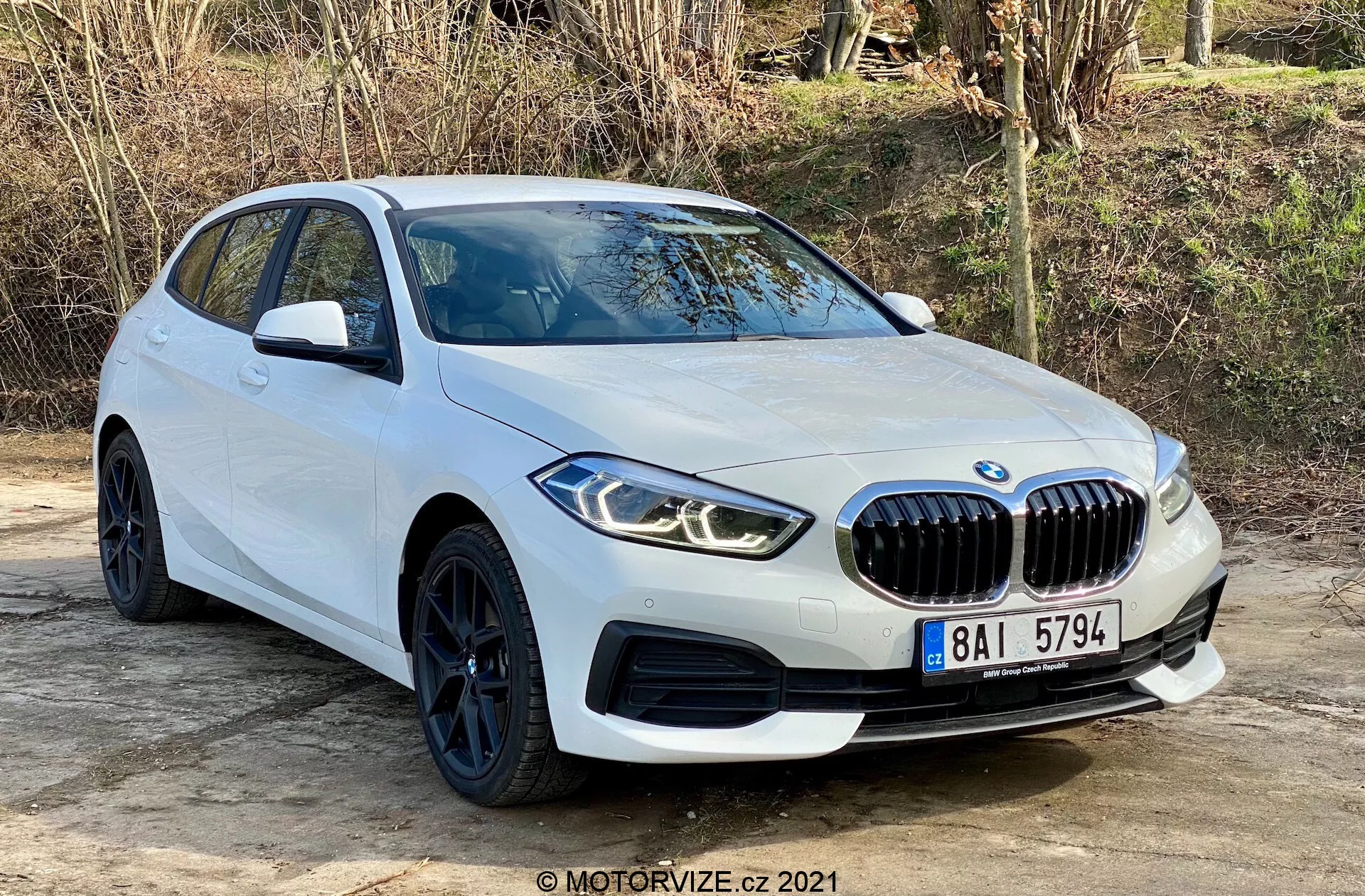 BMW 116i Serie 1 del 2019 en blanco, capturado desde un ángulo de tres cuartos frontal, que muestra su parrilla delantera prominente en forma de riñón, faros LED angulares, elegantes llantas negras con neumáticos de perfil bajo y líneas aerodinámicas del cuerpo. Posicionado al aire libre sobre una superficie plana con vegetación natural en el fondo, la toma resalta el diseño moderno y las características deportivas del coche, incluidos los sensores de estacionamiento y los espejos laterales con señal de giro integrada.