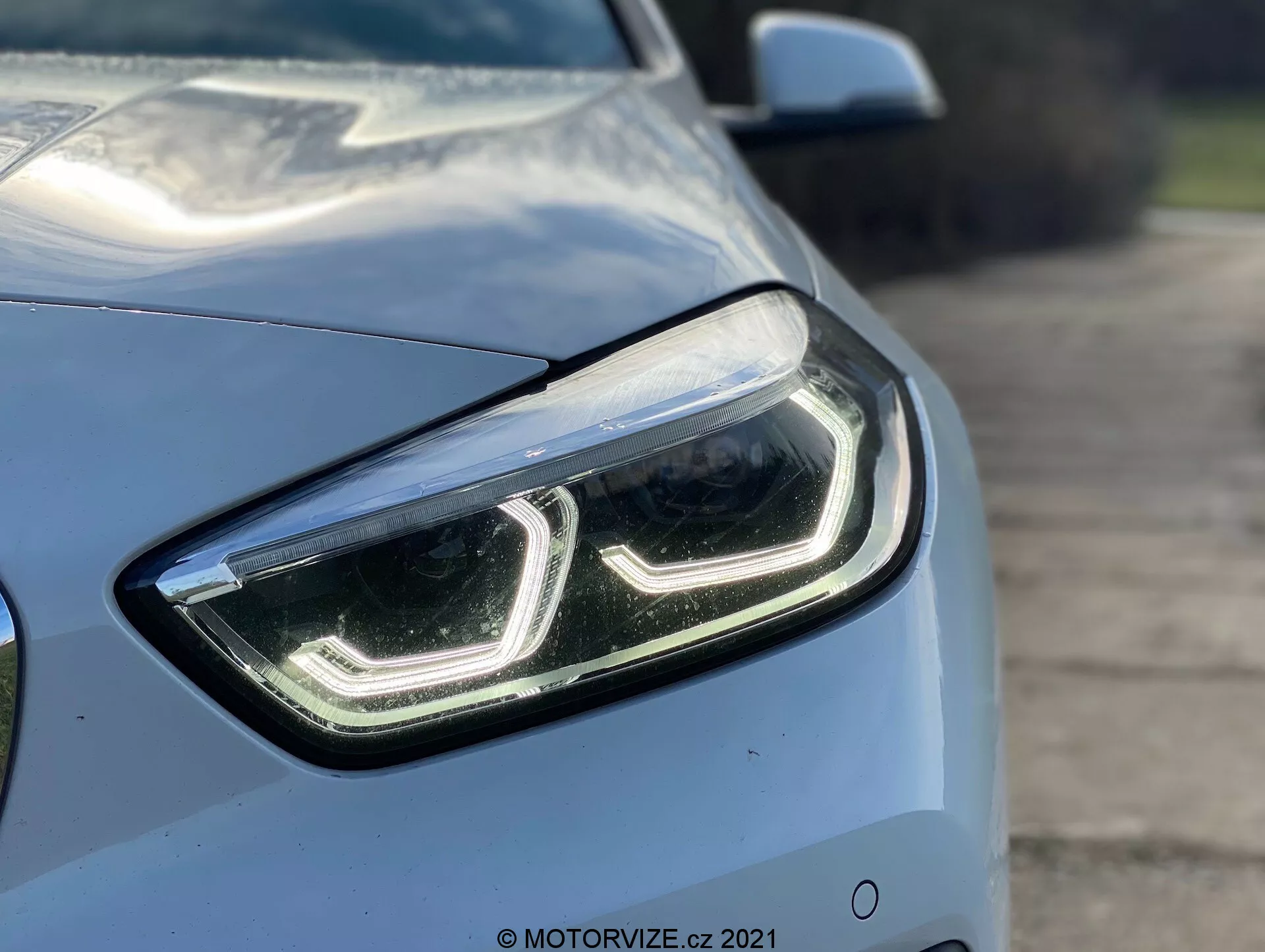 Toma en primer plano del BMW Serie 1 2019 (BMW 116i) desde un ángulo de cuarto delantero, resaltando el lado izquierdo y enfocándose en el diseño moderno y elegante del faro delantero izquierdo con luces diurnas LED y una cubierta de lente clara. Las líneas aerodinámicas del capó y el guardabarros son parcialmente visibles, mostrando el color metálico claro y el acabado brillante del coche. Un fondo difuminado enfatiza las características tecnológicas avanzadas del faro, sugiriendo lámparas LED o HID, posiblemente equipadas con tecnología de iluminación adaptativa.