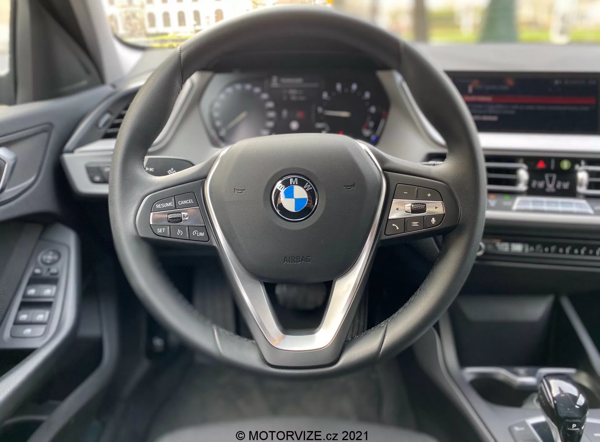 Perspectiva del asiento del conductor de un BMW 1 Series 2019 (BMW-116i), enfocándose en el volante contemporáneo de tres radios con un acento metálico en el radio inferior y el emblema prominente de BMW en el centro. El lado izquierdo del volante incluye controles de medios y comunicación, mientras que el lado derecho tiene botones para el control de crucero y una palanca de cambios visible. Se aprecia un cuadro de instrumentos parcialmente visible con indicadores de estilo analógico y una pantalla de infoentretenimiento en la consola central, que sugiere características avanzadas como navegación GPS y control de clima.