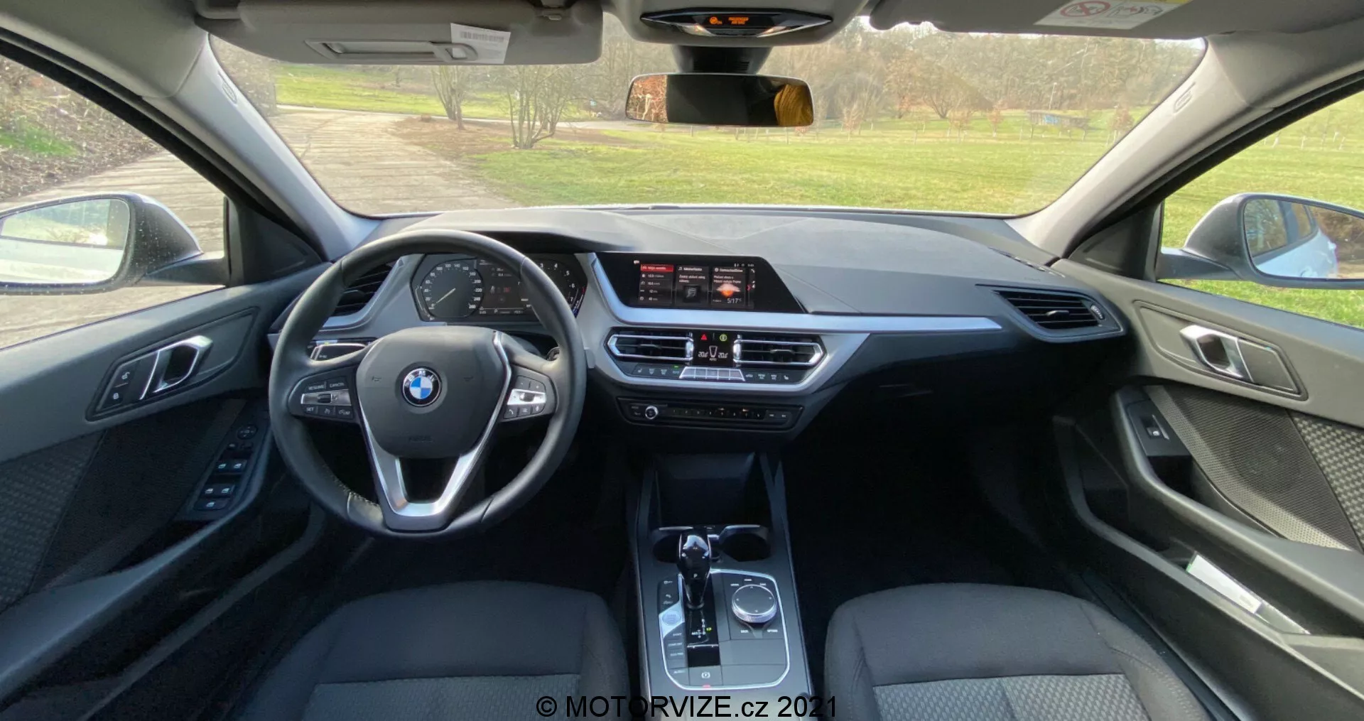 Vista interior desde el asiento trasero de un BMW Serie 1 2019 (BMW-116i), mostrando una cabina moderna con un volante multifunción de tres radios con el logo de BMW, una pantalla de infoentretenimiento independiente, control de clima de doble zona y palanca de cambios automática. El uso de cuero, plásticos suaves y acentos metálicos resalta un nivel de equipamiento de alta gama.
