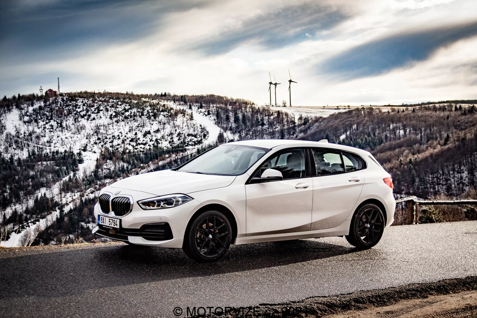 La imagen muestra un hatchback BMW 116i 2019 blanco estacionado en un mirador escénico o camino de montaña, fotografiado desde un ángulo lateral-frontal que muestra la parrilla frontal, los faros LED, la rueda delantera, el perfil lateral y un vistazo a la sección trasera. La perspectiva es ligeramente elevada, destacando el techo del coche, que está desplazado hacia la izquierda, con un fondo de colinas nevadas y turbinas eólicas distantes. El vehículo presenta una parrilla en forma de riñón, neumáticos de perfil bajo en llantas de aleación multirradio, y llantas de colores oscuros, espejos laterales con señales de giro integradas y ventanas tintadas. El entorno sugiere un clima claro, mejorando la naturaleza promocional y visualmente atractiva de la fotografía, potencialmente tomada durante un viaje por carretera o una revisión automotriz.
