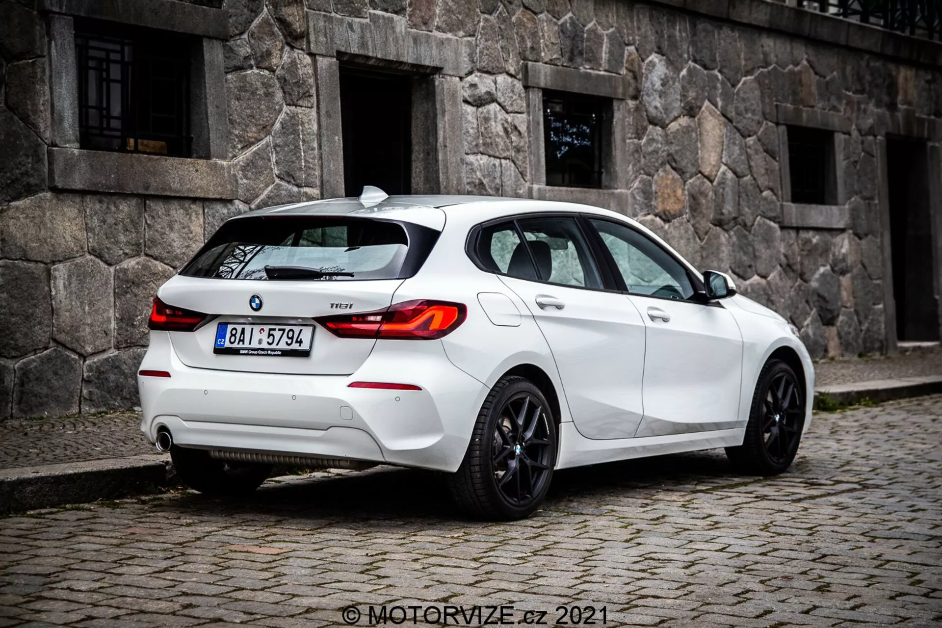 Imagen a media distancia a nivel del suelo de un hatchback blanco BMW 1er-series 116i 2019 capturada desde un ángulo trasero de tres cuartos, mostrando prominentemente las luces traseras con diseño LED, el parabrisas trasero y llantas deportivas. Las características visibles incluyen sensores en el parachoques trasero posiblemente para asistencia de estacionamiento, una pequeña antena en el techo probablemente para GPS o radio satelital, insignia del modelo en el maletero, sistema de escape dual que sugiere rendimiento, y ventanas tintadas para privacidad o confort.