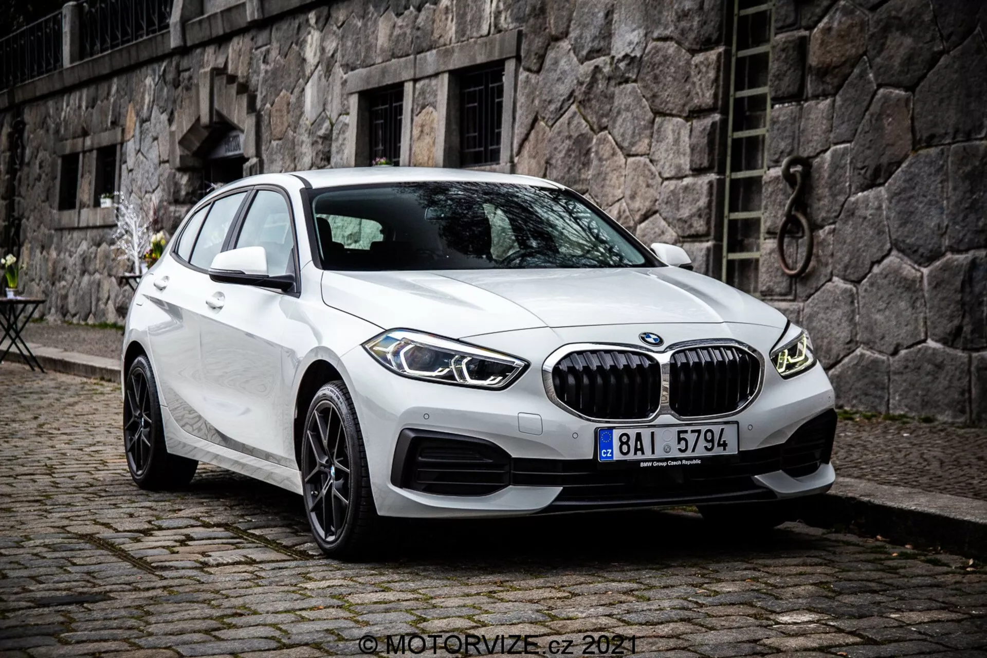 La imagen muestra un BMW Serie 1 de 2019, específicamente el BMW 116i, en color blanco. Fotografiado desde un ángulo de tres cuartos frontal, muestra la fachada delantera del coche, el lado del pasajero y el diseño detallado de sus ruedas. La foto se toma desde un punto de vista ligeramente elevado, sugiriendo que el fotógrafo estaba de pie o usando un trípode. El coche está estacionado con las ruedas delanteras giradas a la derecha, revelando una vista clara de las llantas y el perfil lateral del neumático. Las características incluyen una parrilla frontal prominente con lamas verticales, líneas esculpidas en el capó y los paneles laterales, faros LED delgados y entradas de aire en el parachoques que podrían alojar sensores de aparcamiento. Los espejos laterales con señales de giro integradas y las ventanas sin marco destacan su estética premium. Las llantas de aleación de múltiples radios sugieren un nivel de equipamiento superior o una actualización opcional. La ventana trasera empinada enfatiza su perfil de hatchback. El vehículo está estacionado sobre un pavimento de adoquines en un entorno urbano o histórico con un edificio de paredes de piedra y una pequeña mesa de estilo bistró en el fondo. La imagen es nítida y detallada, capturada con luz diurna natural y nubosa.