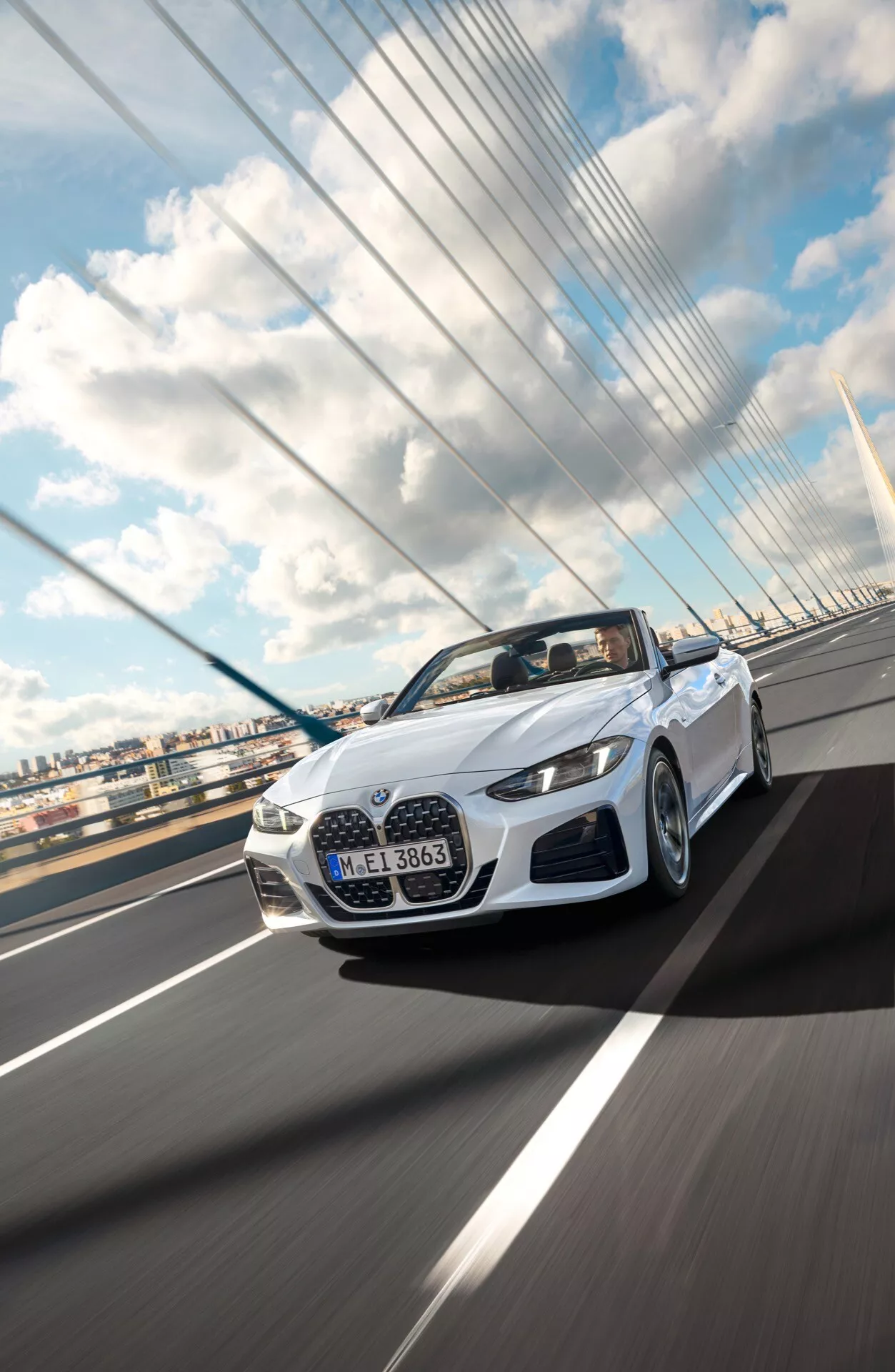 Convertible BMW serie 4 del 2024 (facelift G22-G23-G26-G82-G83) en blanco, captado dinámicamente desde un ángulo frontal bajo mientras conduce en un puente colgante bajo un cielo parcialmente nublado, mostrando su elegante diseño, iluminación LED y características aerodinámicas
