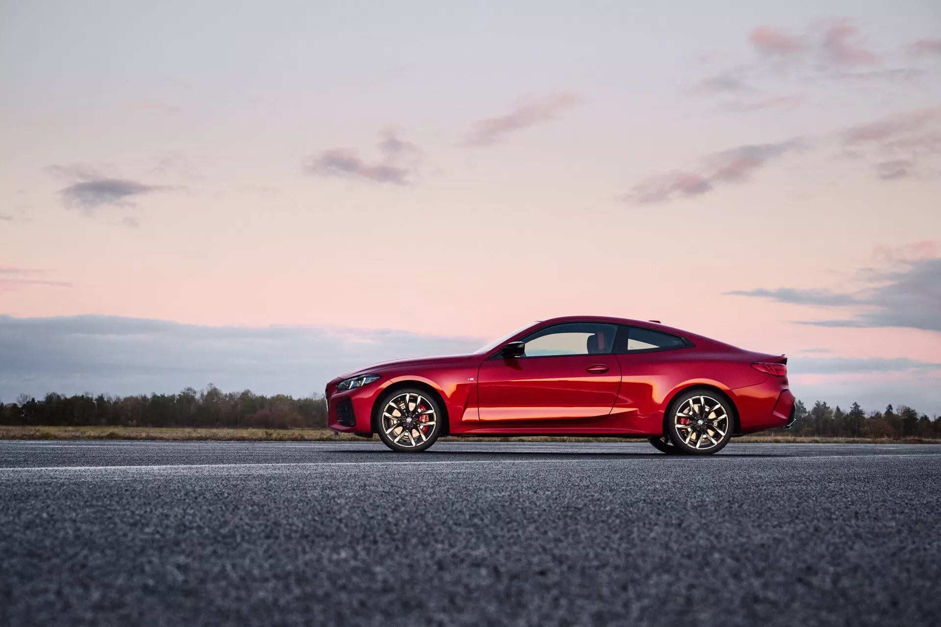 Deportivo coupé rojo BMW Serie 4 (G22-G23-G26-G82-G83 Facelift) de dos puertas en pista de aterrizaje con cielo crepuscular de fondo, destacando diseño elegante, aerodinámico con grandes ruedas de aleación de múltiples radios y características modernas.