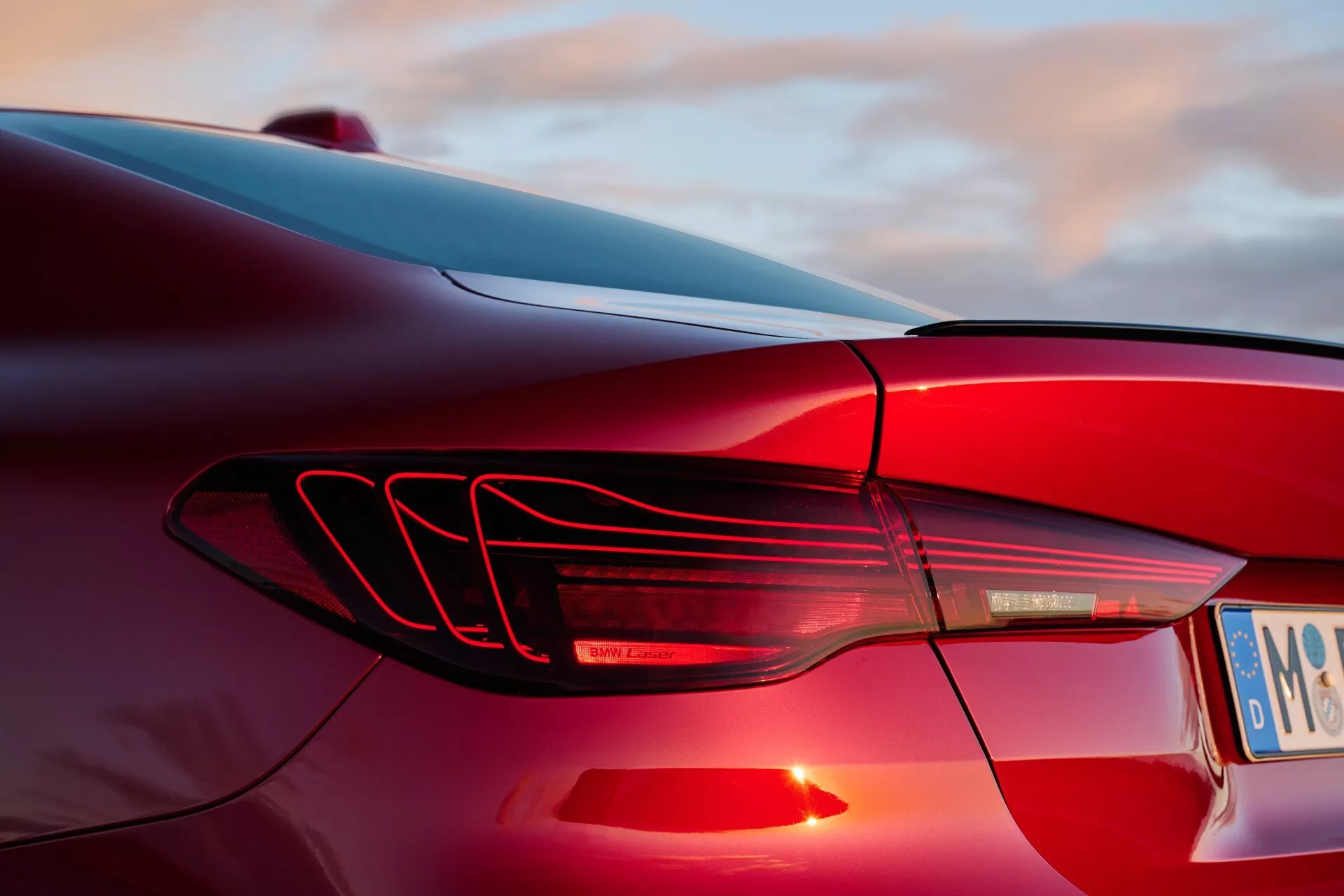 Primer plano de la luz trasera izquierda del BMW Serie 4 2024 (G22-G23-G26-G82-G83 lifting) con tiras de luces LED horizontales y diseño de aletas aerodinámicas, contra un fondo de cielo al atardecer