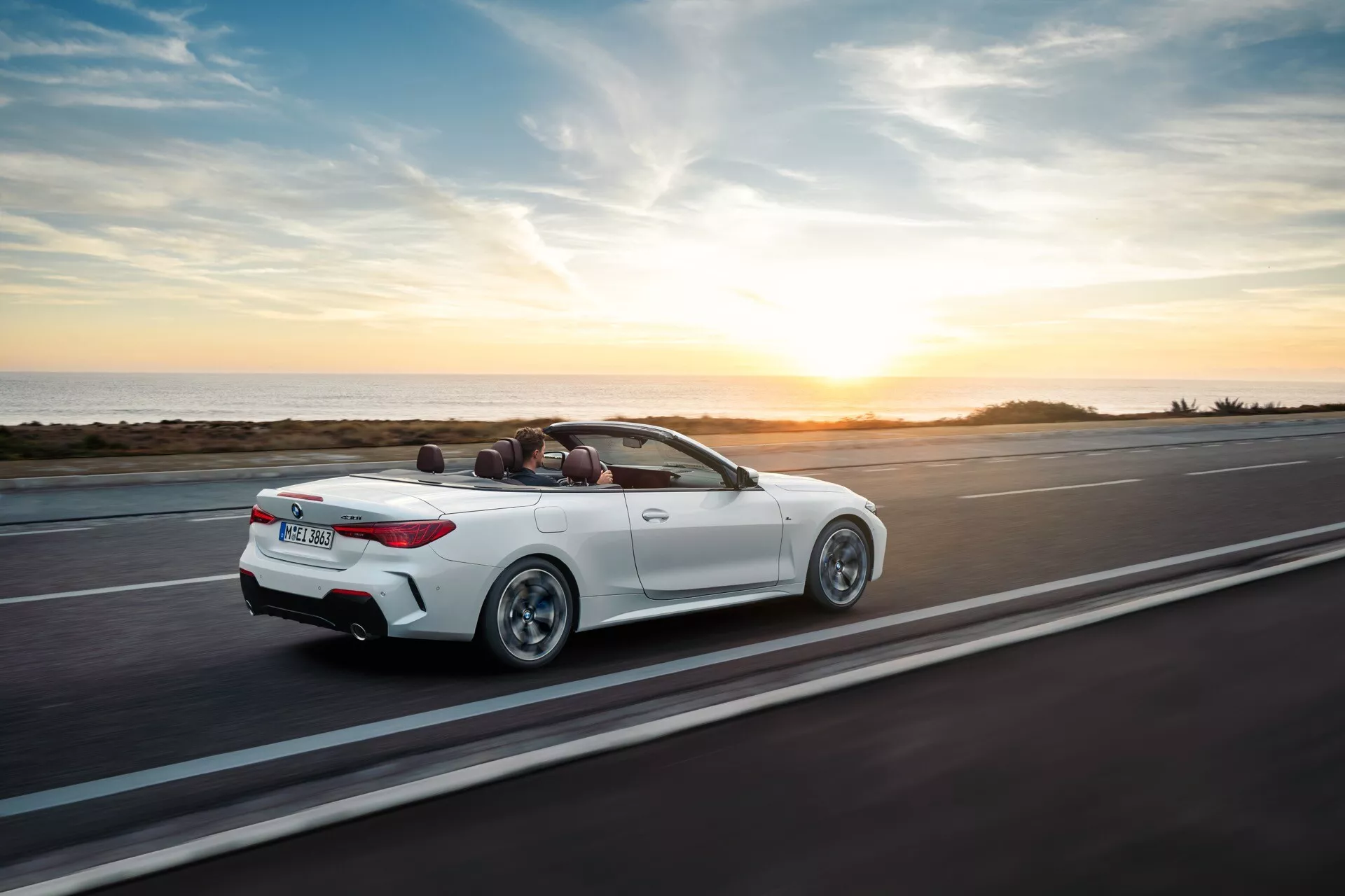 BMW Serie 4 Convertible (G22-G23-G26-G82-G83 rediseñado) en movimiento en una carretera costera al atardecer, vista trasera aérea de tres cuartos que muestra su exterior blanco, capota convertible bajada y diseño deportivo