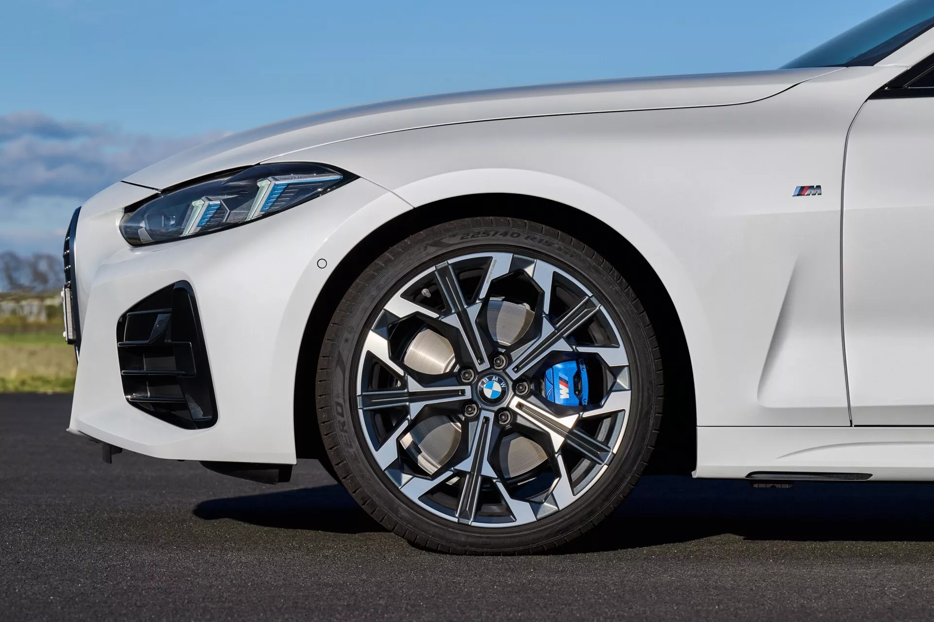 Vista lateral de cerca del BMW Serie 4 2024 (G22-G23-G26-G82-G83 Facelift) en blanco, centrada en la rueda delantera con llanta de aleación de múltiples radios, pinza de freno azul y entradas de aire orientadas al rendimiento bajo la luz natural del día