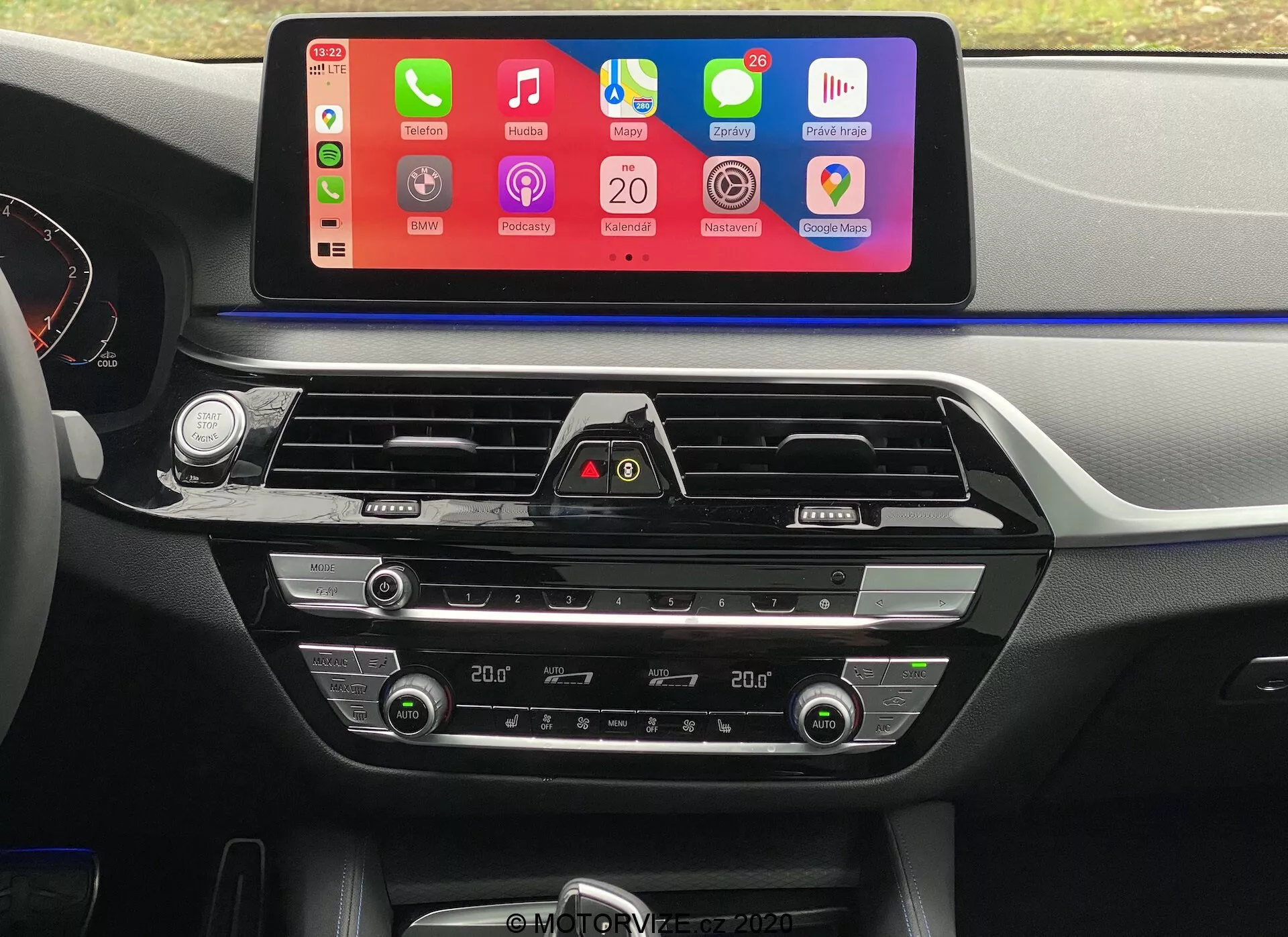 Toma interior de un BMW 5er-Reihe 2020 (BMW 520i Touring Steptronic) desde la perspectiva del asiento del conductor, enfocándose en el tablero central. La característica más destacada es una pantalla táctil de infoentretenimiento grande y horizontal con una interfaz colorida que permite la integración de smartphones como Apple CarPlay y Android Auto. Debajo de la pantalla se encuentran salidas de aire con detalles cromados y un botón de luces de emergencia. Más abajo están los controles de climatización de doble zona con pantallas digitales para ajustar la temperatura, botones y diales para la temperatura del aire, velocidad del ventilador y distribución del aire. El tablero y la consola central están hechos de plástico suave al tacto y acentos metálicos, aumentando el atractivo estético.