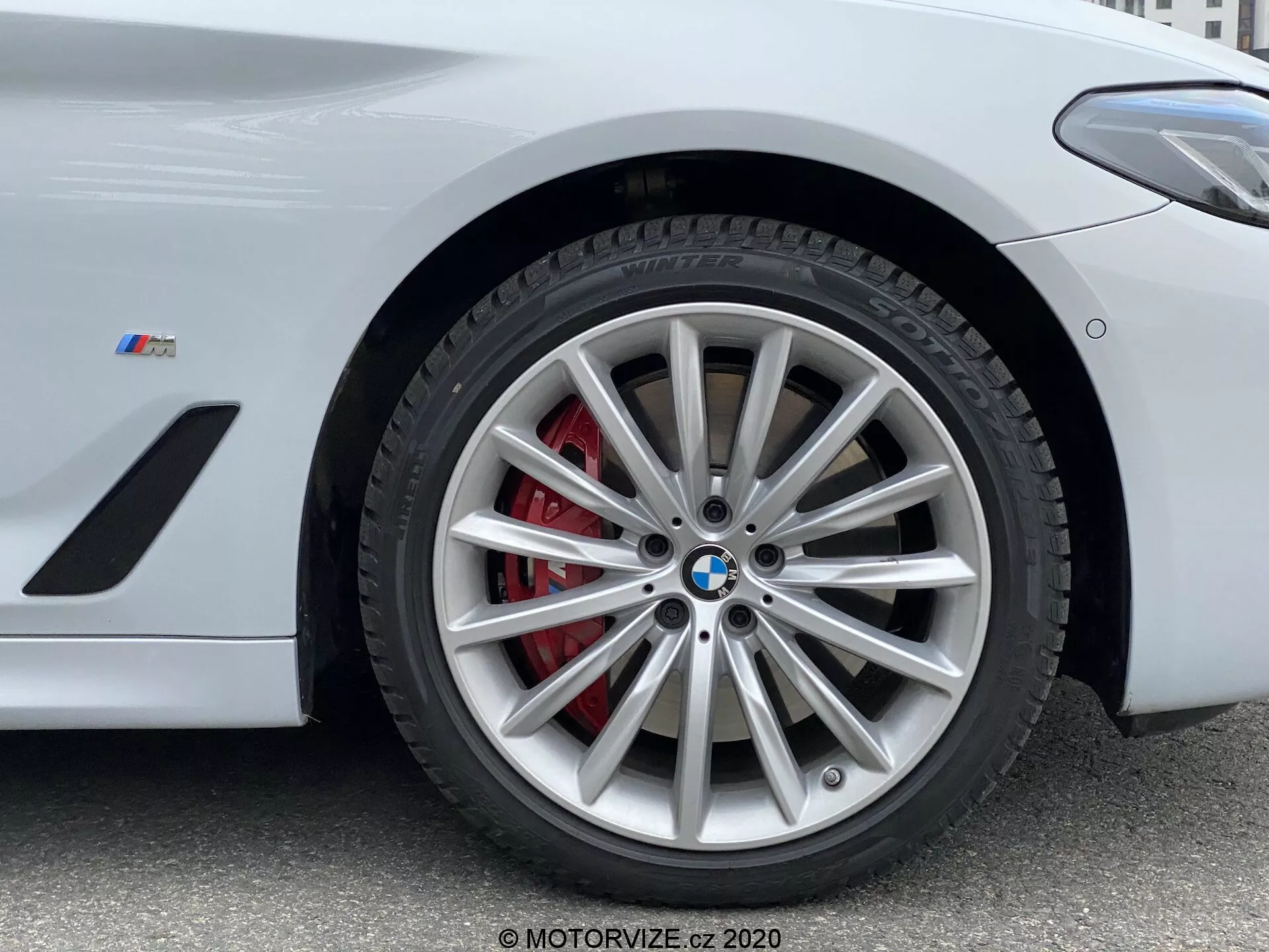 Vista de primer plano de la sección de la rueda delantera de un BMW de la Serie 5 2020, específicamente el modelo 520i Touring Steptronic. La fotografía en ángulo bajo resalta el intrincado diseño del aleación de múltiples radios con acabado plateado, complementado por una pinza de freno roja y un rotor ventilado. El emblema de BMW y la insignia 'M' en el guardabarros señalan que este es un vehículo orientado al rendimiento de la serie M de BMW. La imagen captura detalles clave adecuados para mostrar sistemas de frenos de alto rendimiento, diseño de vehículos de lujo y tecnología de neumáticos.