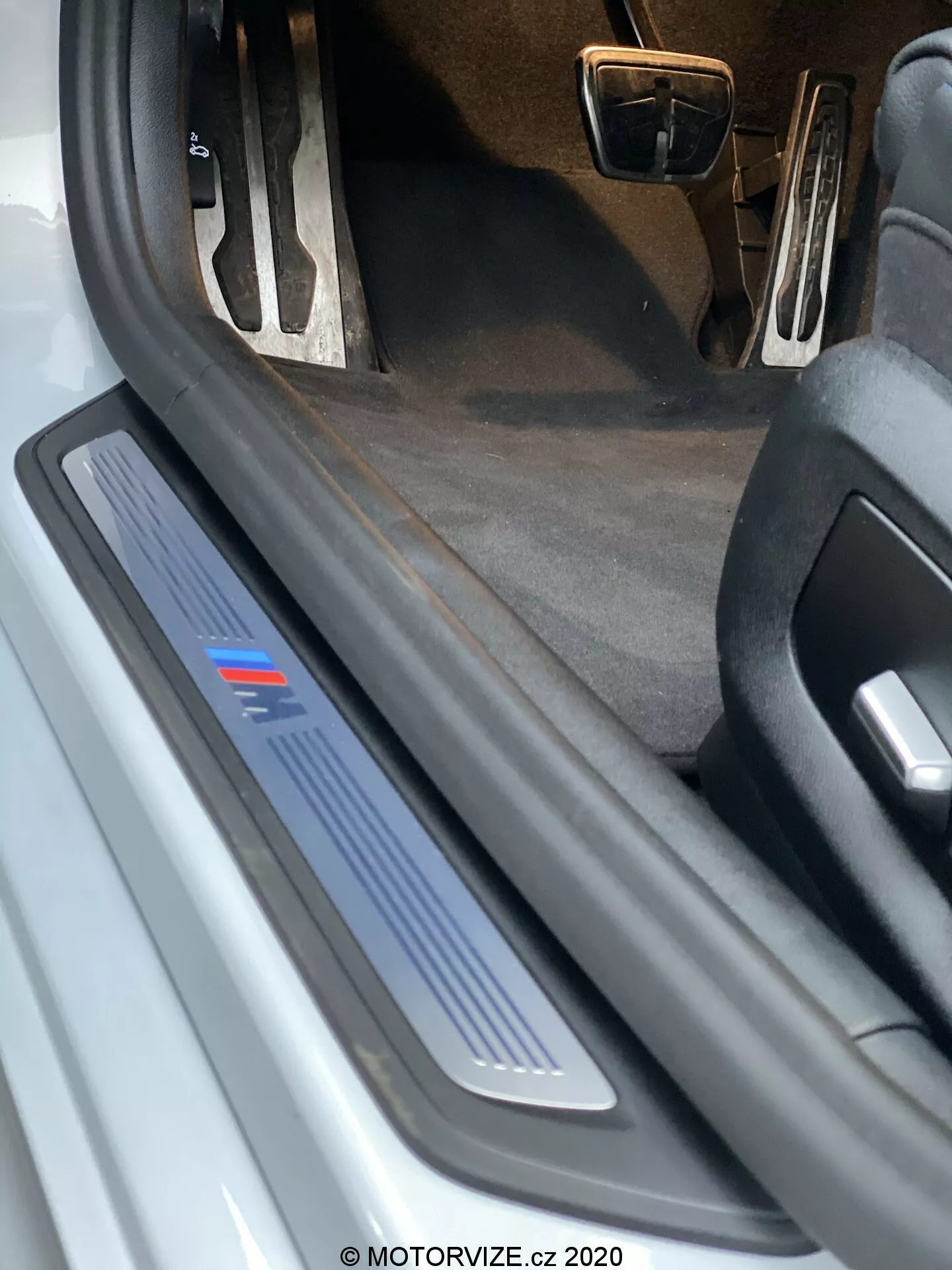 Vista en primer plano del umbral de la puerta del lado del conductor de un BMW 520i Touring Steptronic 2020. La imagen muestra una perspectiva a baja altura, resaltando la placa del umbral con acabado metálico y una franja azul, que podría indicar un nivel de equipamiento específico o edición deportiva. No se ve la puerta abierta, pero es evidente la placa del umbral protectora y decorativa, lo que sugiere posible iluminación LED. El interior oscuro de la alfombra y la vista parcial de los pedales con acabado metálico indican un posible paquete interior deportivo. Hay residuos y desgaste visible en la placa del umbral, lo que sugiere que el automóvil está usado.
