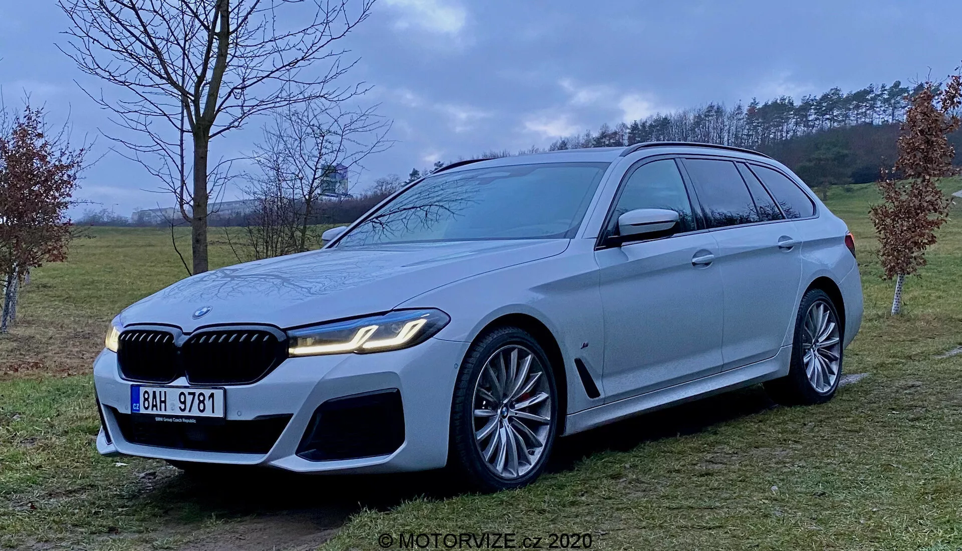 Imagen del BMW Serie 5 Touring 2020 (BMW 520i Touring Steptronic), un coche familiar de lujo en color plateado claro o gris, que muestra su parte delantera agresiva y deportiva con grandes entradas de aire, una parrilla prominente y faros LED elegantes. El vehículo presenta grandes llantas de aleación con diseño de múltiples radios y notables pinzas de freno, una línea de techo larga con spoiler, molduras cromadas en las ventanas y una fuerte línea de hombro. Fotografiado desde la altura del medio del coche en un ambiente rural con luz difusa.