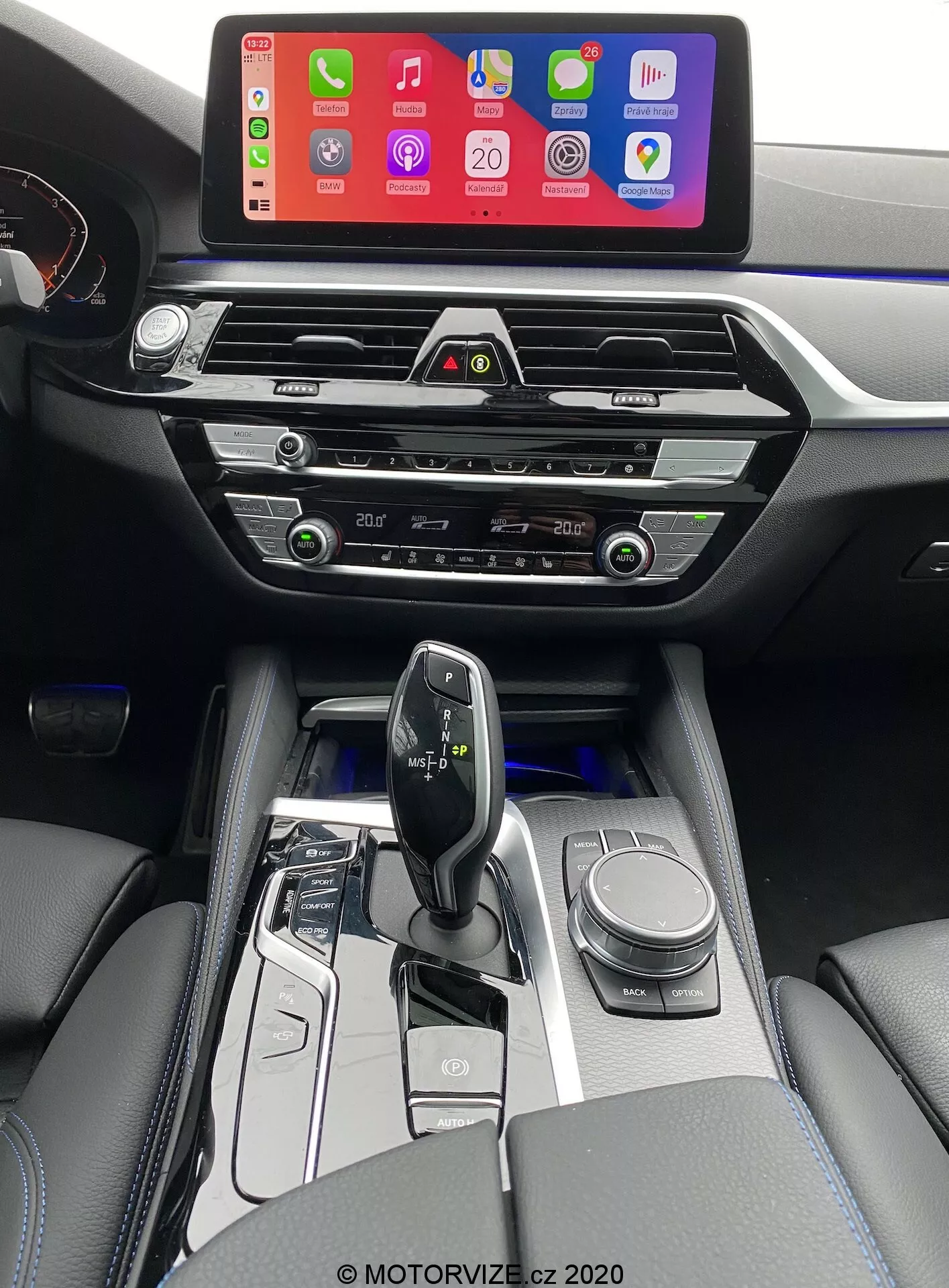 Imagen del interior de un BMW 520i Touring 2020 con transmisión Steptronic, tomada desde la perspectiva del conductor y ligeramente elevada para dar una vista más clara de la consola central, el área de la palanca de cambios y parte del tablero. La imagen muestra una gran pantalla de infoentretenimiento que muestra una interfaz de conexión para teléfonos inteligentes, probablemente Apple CarPlay, montada en lo alto del tablero para una fácil visibilidad. Debajo de la pantalla se encuentran las ventilaciones de aire horizontales modernas con acentos cromados, seguidas de botones y diales táctiles para el control del clima. La consola central también alberga los controles del sistema de audio, controles adicionales de clima y una posible ranura para reproductor de CD. La palanca de cambios ergonómica y minimalista, probablemente electrónica, está rodeada de adornos en negro brillante y metálicos, con botones para la selección del modo de conducción y un dial giratorio para la navegación. El interior también incluye reposabrazos tapizados en cuero con costuras visibles, lo que añade un toque de lujo. La iluminación blanca suave resalta los botones alrededor del área de la palanca de cambios, indicando una transmisión automática.