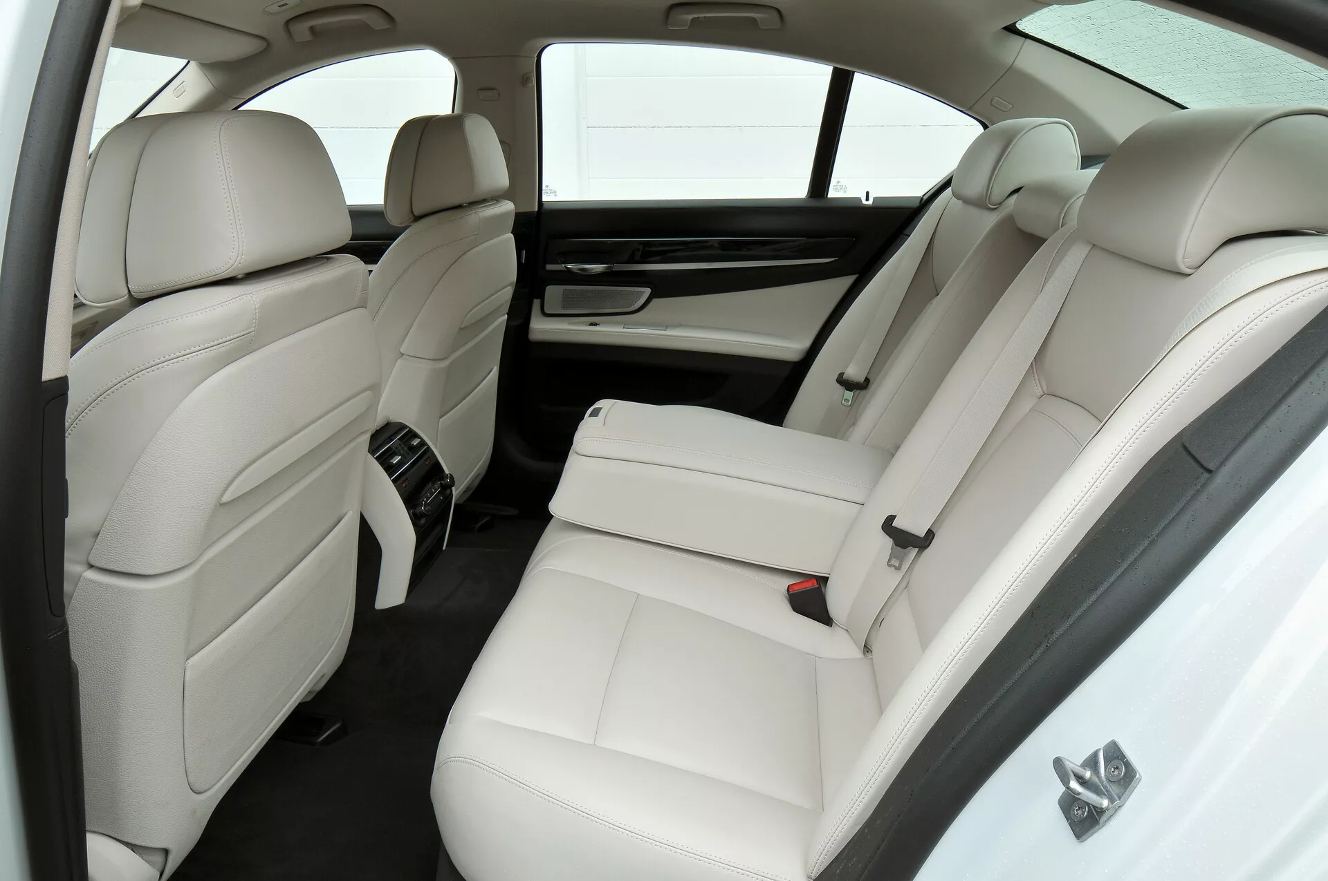 La imagen muestra el lujoso interior de cuero blanco de un BMW Serie 7 (2012 con facelift, F01-F02) enfocándose en el compartimento trasero del pasajero desde una perspectiva ligeramente elevada. Presenta dos asientos traseros separados con reposacabezas y refuerzos laterales, una consola central con controles, ventilas de control climático traseras, una cortina solar en la ventana trasera, reposacabezas integrados en los respaldos de los asientos delanteros y una paleta de colores minimalista en blanco y tonos oscuros.