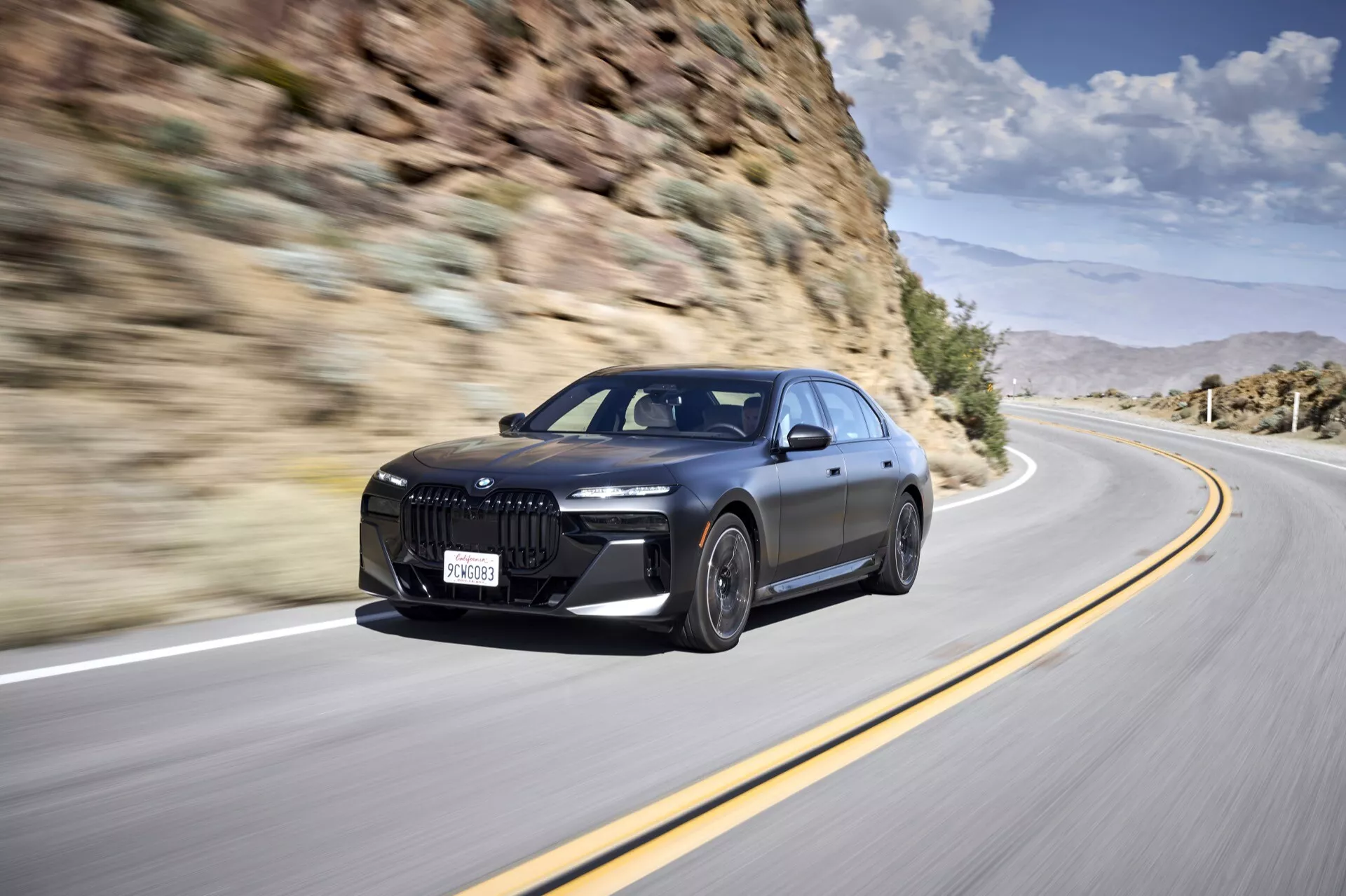 Sedán de lujo BMW i7 (G70) 2022 en movimiento, destacando su gran parrilla frontal distintiva, faros LED, parachoques frontal aerodinámico con grandes entradas de aire, líneas deportivas en el capó, grandes ruedas de aleación y perfil lateral elegante con manijas de puerta al ras, contra un fondo montañoso.