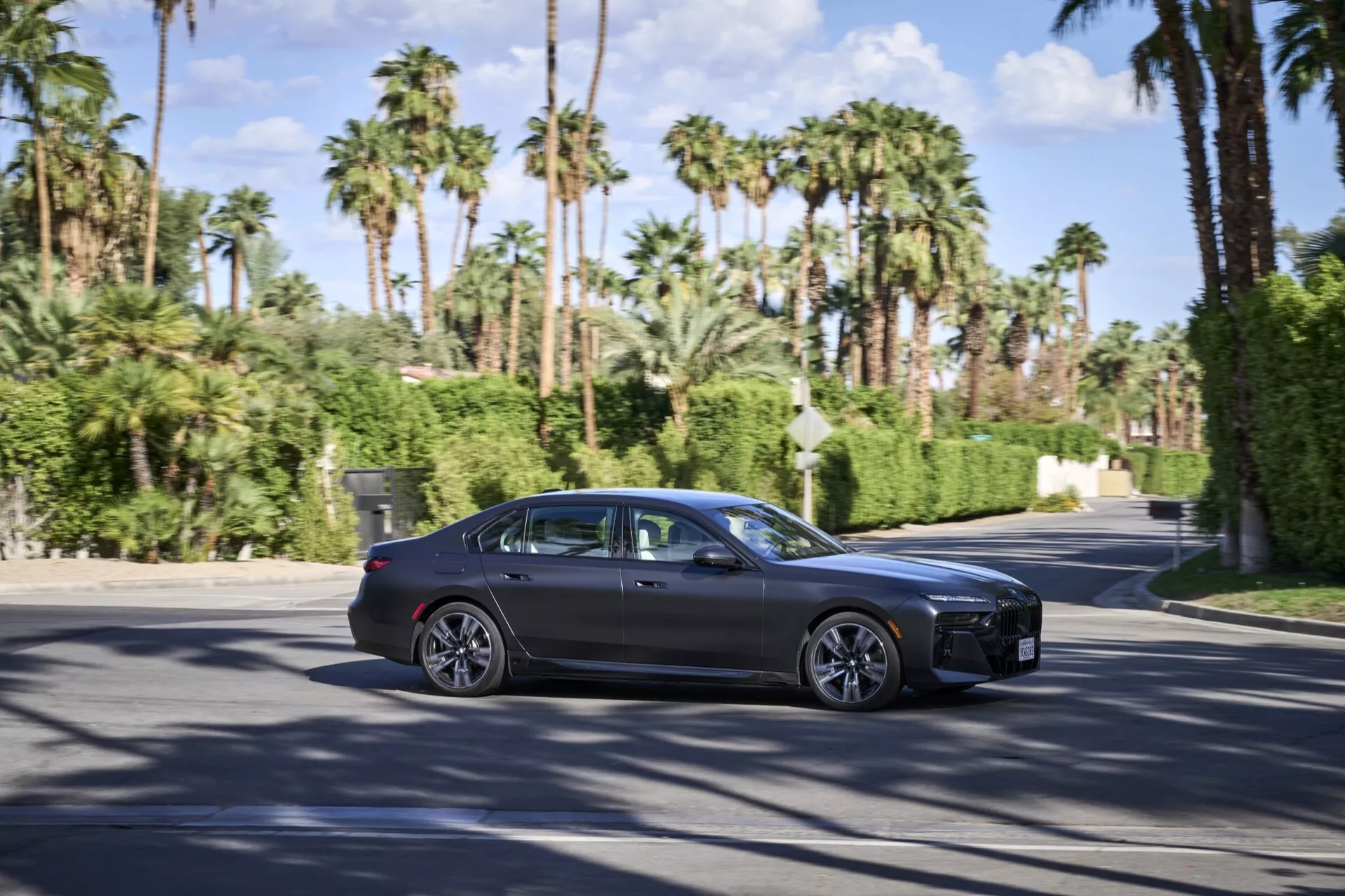 Sedán de cuatro puertas de lujo BMW i7 (G70) de 2022 en movimiento en una carretera soleada con palmeras, mostrando grandes ruedas de aleación, acentos de cromo en las ventanas, espejos laterales del color de la carrocería y ventanas tintadas