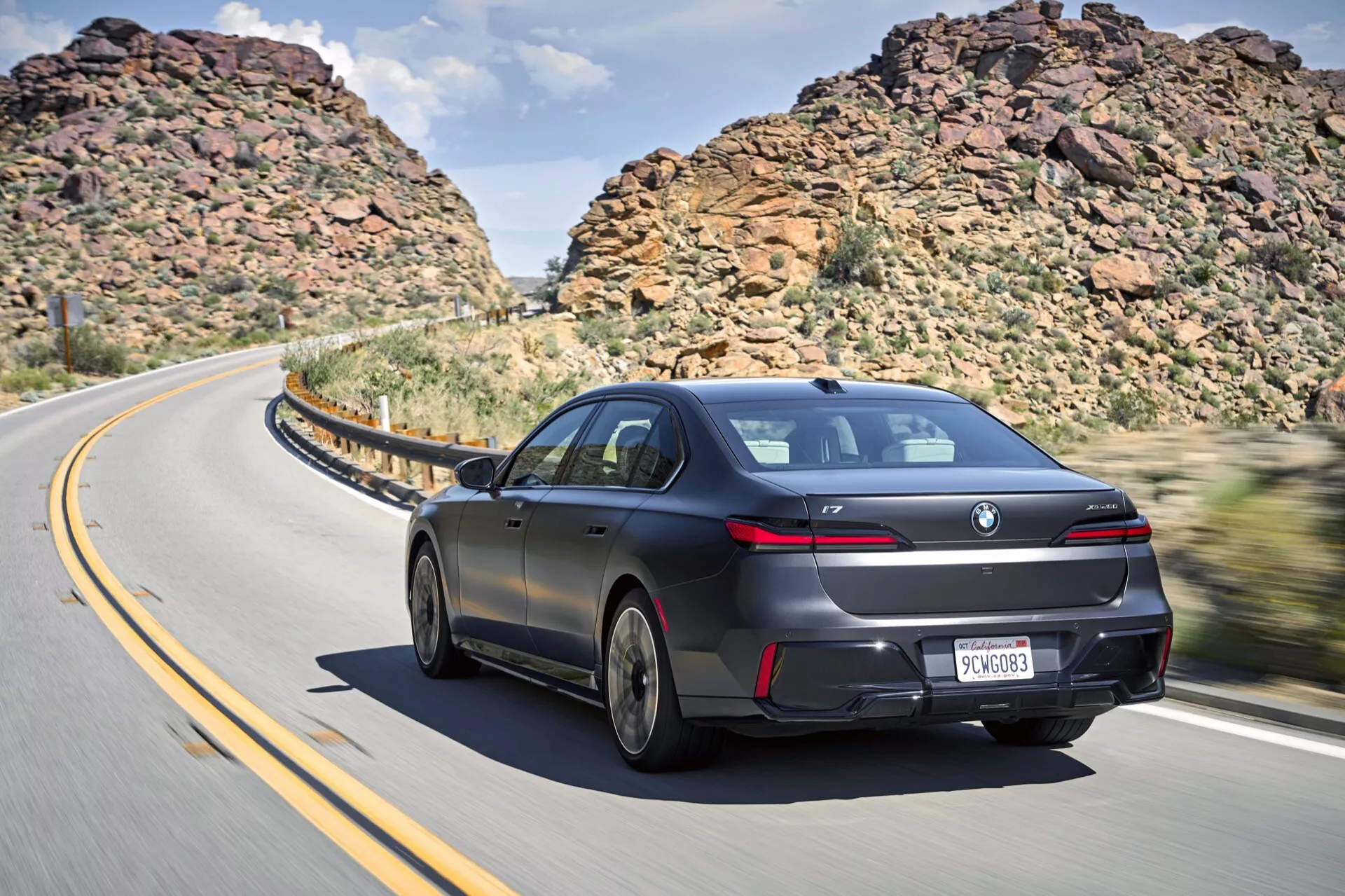 Vista trasera dinámica de tres cuartos de un sedán BMW i7 2022 (G70) en movimiento sobre una carretera curva con laderas accidentadas y un cielo parcialmente nublado, presentando un diseño aerodinámico elegante, pintura lujosa, ruedas de aleación de diseñador, luces traseras LED, spoiler trasero sutil, ventanas tintadas y salidas de escape dobles.