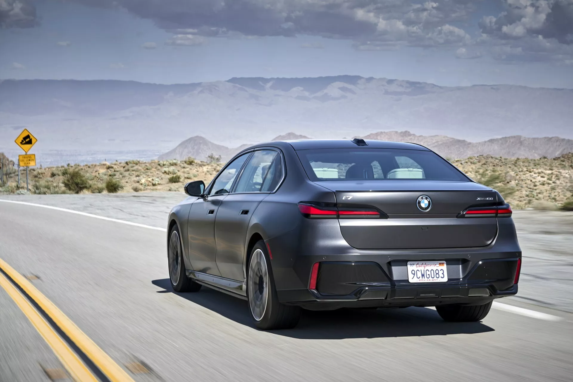 Sedán moderno BMW i7 (G70) de 2022 en acabado mate, capturado en movimiento desde un ángulo trasero externo y de tres cuartos en una carretera del desierto, mostrando su diseño aerodinámico, luces traseras LED y características tecnológicas como sensores de estacionamiento y techo solar, bajo un cielo parcialmente nublado.