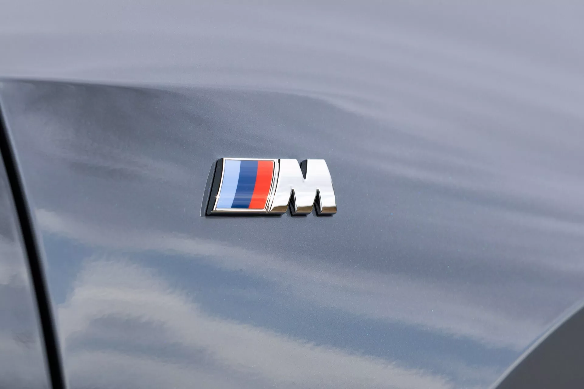 Vista de cerca del emblema de la serie M de BMW en el maletero de un BMW X1 (U11) 2022 con acabado metálico gris oscuro, mostrando el emblema rectangular con fondo negro y rayas azules y rojas seguido de la letra blanca 'M'