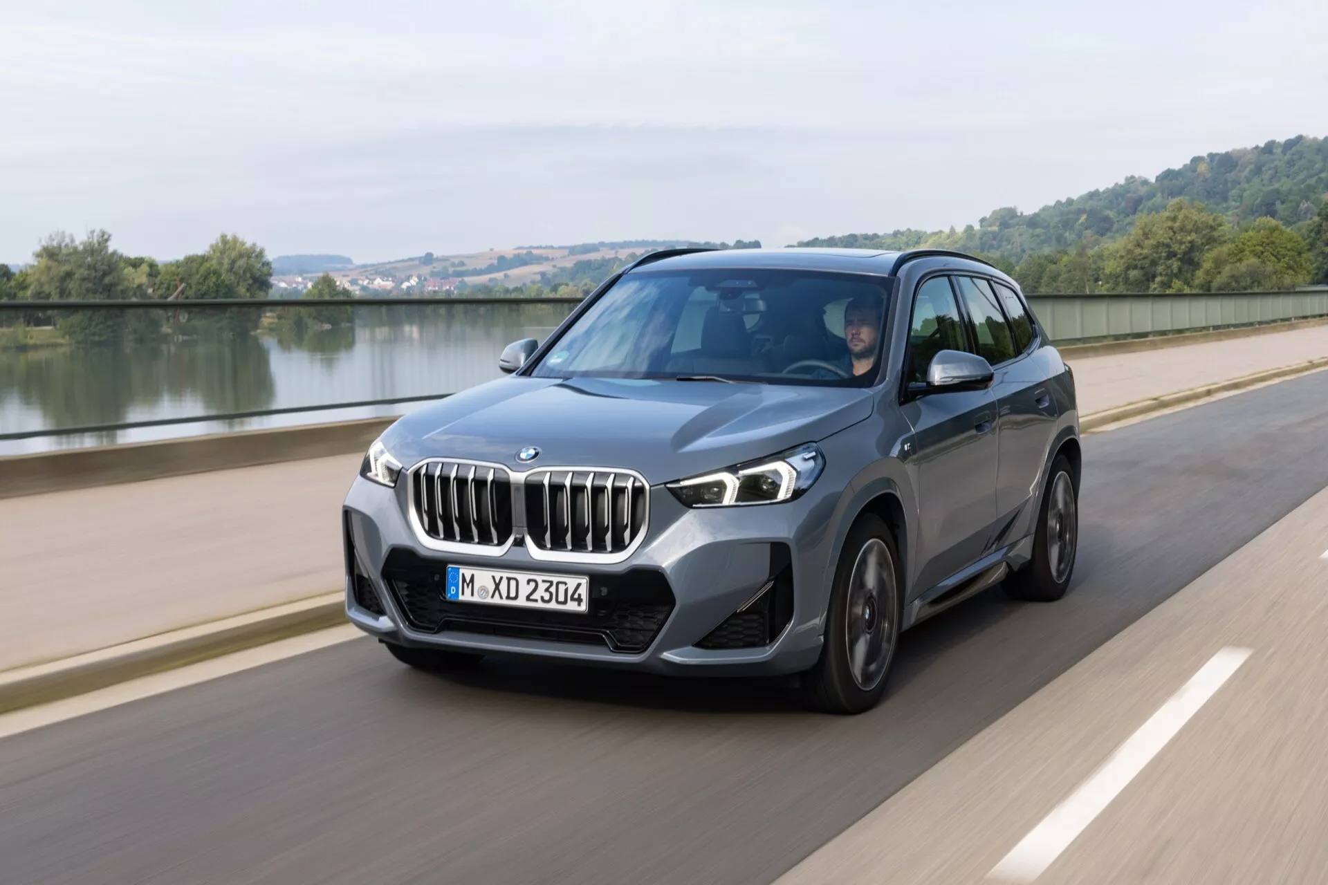 SUV crossover de lujo BMW X1 (U11) 2022 en movimiento por una carretera suave junto a un cuerpo de agua, destacando diseño dinámico, faros LED y funciones tecnológicas avanzadas