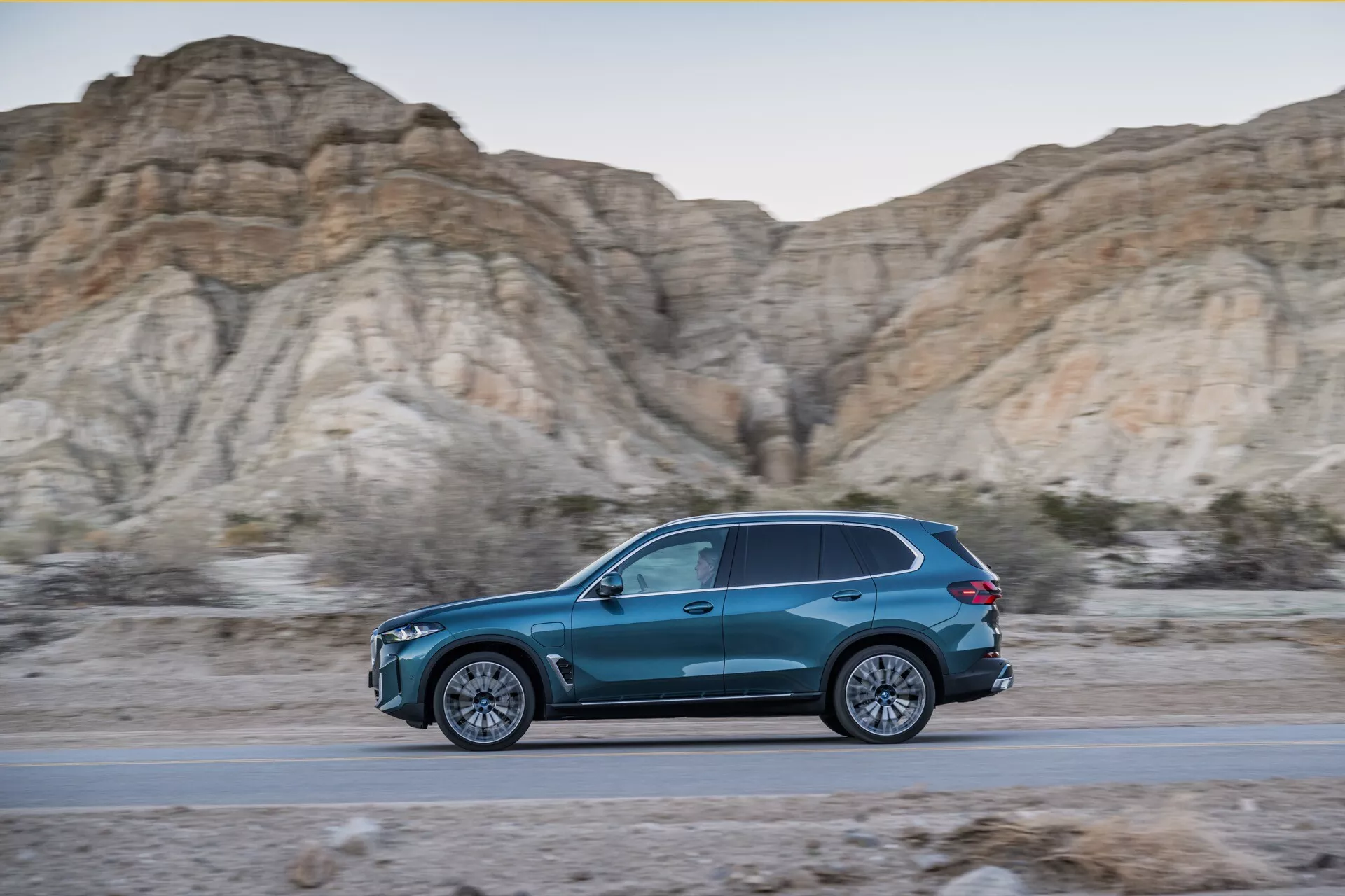 Perfil lateral dinámico del BMW X5 2023 (G05-F95 facelift) en movimiento, SUV azul con forma de lágrima en una carretera con fondo de colinas áridas y vegetación escasa durante la hora dorada, mostrando un diseño elegante, eficiencia aerodinámica, ruedas de aleación de múltiples radios y iluminación LED