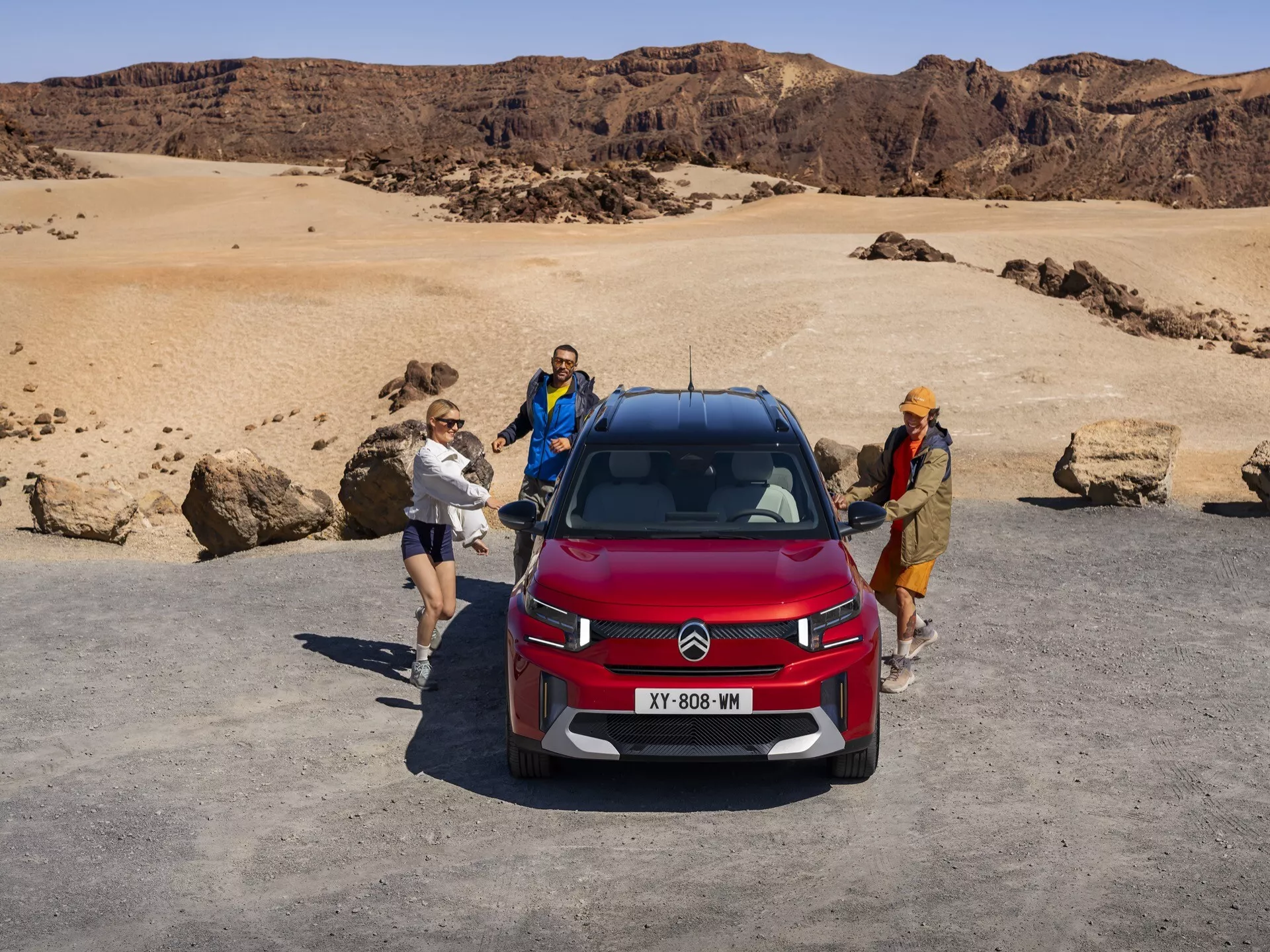 Vista frontal alta de un SUV Citroën C5 Aircross rojo que muestra el capó, la parrilla con el distintivo logo de doble chevron y parte del techo. Destaca el diseño de los faros LED de dos niveles, el módulo de radar o sensor integrado para sistemas avanzados de asistencia al conductor y el revestimiento robusto de los arcos de rueda, ubicado en un terreno similar a un desierto con tres personas interactuando alrededor del coche.
