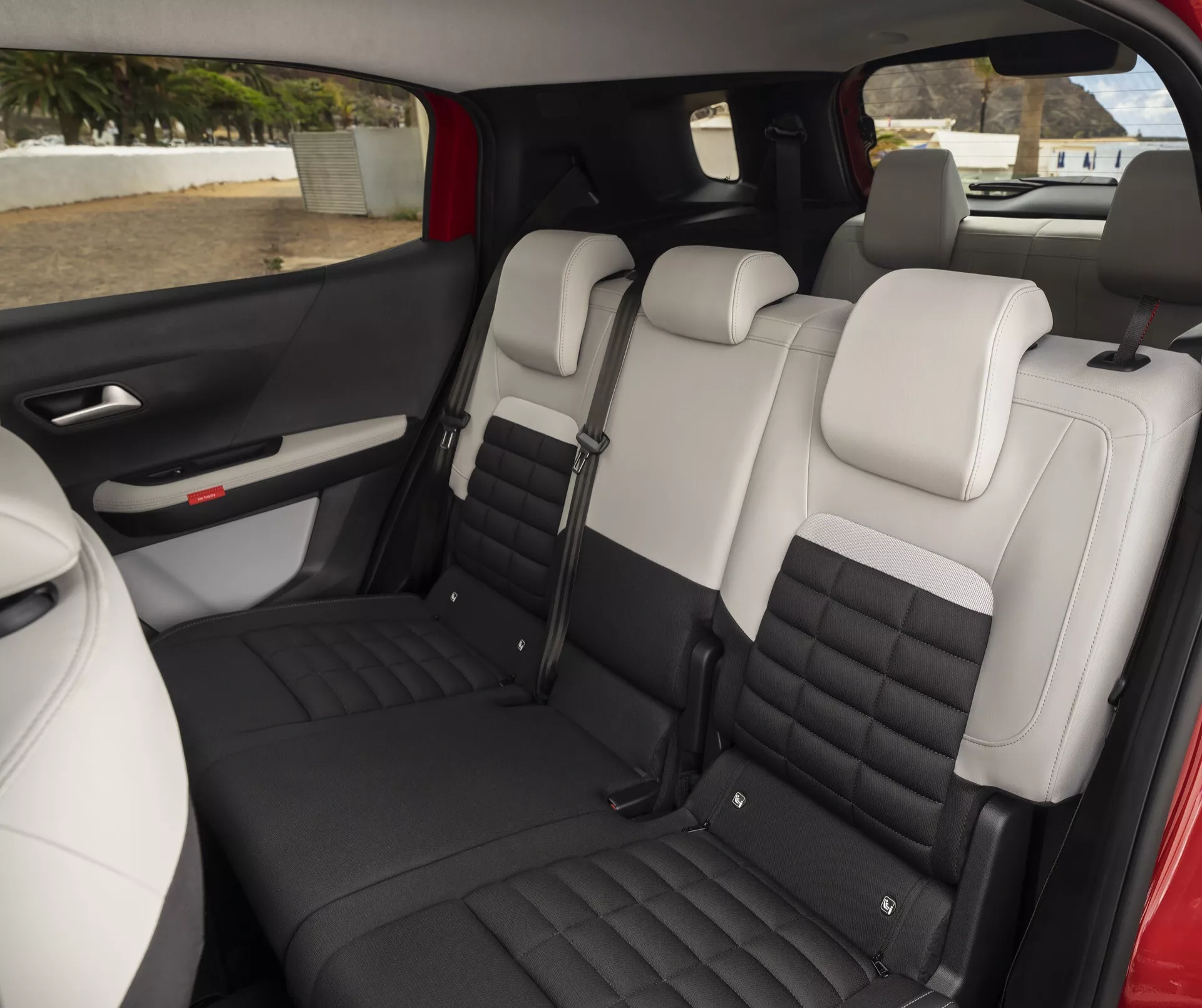 La imagen muestra el interior del Citroen C3 Aircross 2024 (2ª generación) centrándose en la zona de pasajeros trasera. Vista desde el lateral, la perspectiva es ligeramente superior al nivel de los asientos, probablemente tomada desde el área del asiento del pasajero delantero o desde la puerta lateral abierta a la altura de pie. El asiento trasero tipo banco puede acomodar a tres pasajeros y cuenta con tapicería de dos tonos, con partes centrales en crema o blanco y bordes en negro o gris oscuro. Los asientos presentan un diseño acolchado con líneas de costura horizontales, enfatizando la comodidad y el estilo. Hay un apoyabrazos plegable con portavasos o compartimento de almacenamiento integrado, indicando conveniencia y un posible asiento dividido 60/40. El panel de la puerta lateral muestra un interruptor de ventana eléctrica, una manija de puerta y la rejilla del altavoz, destacando características modernas. La gran ventana trasera, posiblemente tintada, sugiere una cabina espaciosa con una sensación abierta y aireada. El diseño del interior, los materiales y el esquema de colores implican un vehículo moderno de gama media a alta enfocado en la comodidad.