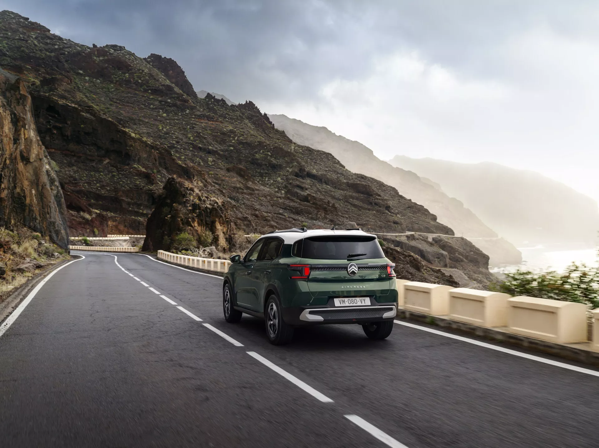 Vista trasera tres cuartos del Citroen C3-Aircross 2024 (2ª generación) mostrando luces traseras LED, insignias en el maletero, líneas de desempañador en el parabrisas trasero, llantas de aleación y un parachoques trasero sin fisuras, posiblemente con sensores de estacionamiento. El coche está en una carretera curva costera o montañosa, mejorando su atractivo para viajes y ocio.
