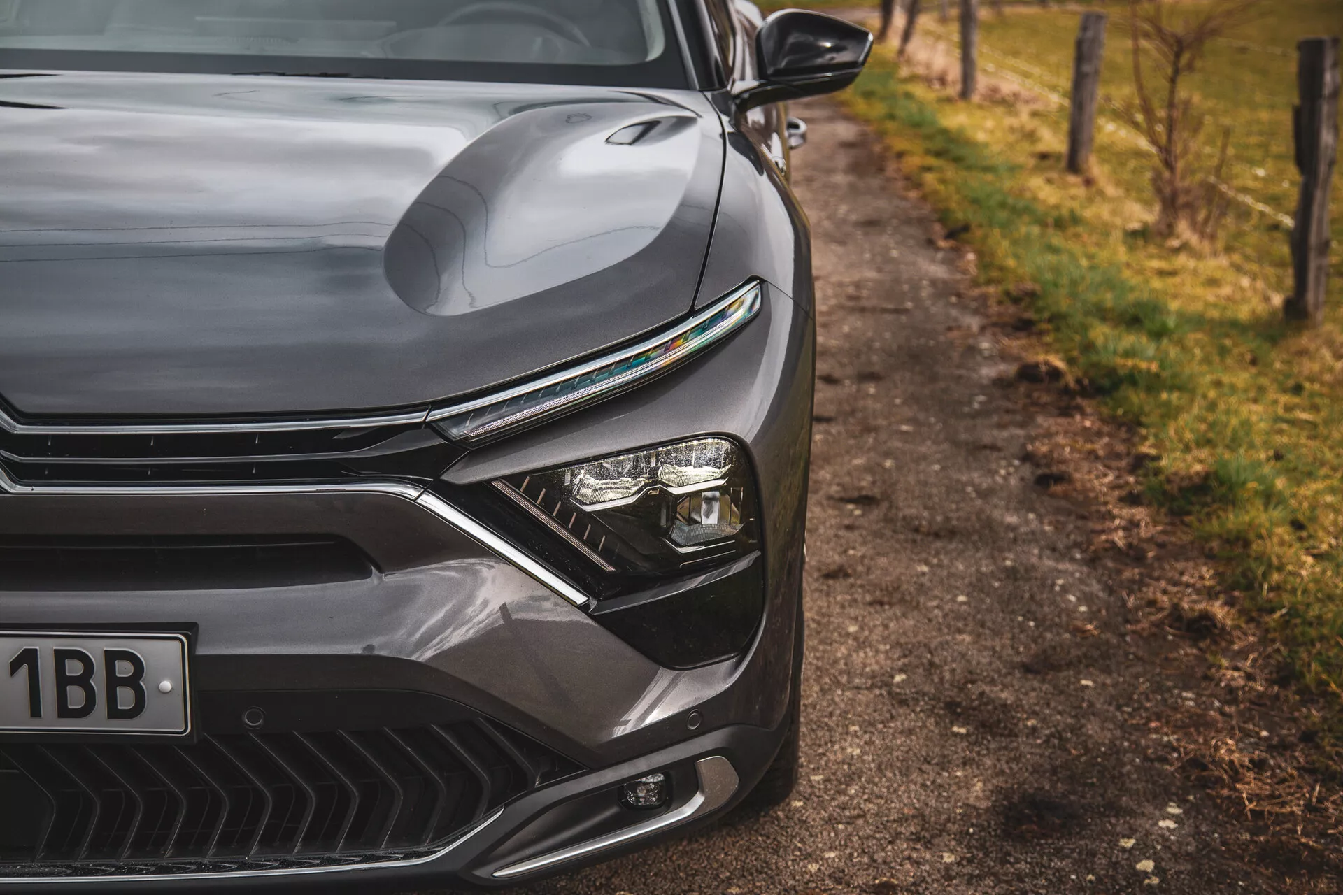 Primer plano del Citroen C5-X 2022 mostrando el elegante diseño del lado frontal izquierdo, incluyendo el capó contorneado, la parrilla pronunciada con líneas horizontales, el avanzado sistema de faros LED y el paragolpes frontal aerodinámicamente formado con tomas de aire, situado contra un fondo natural desenfocado al borde de la carretera.