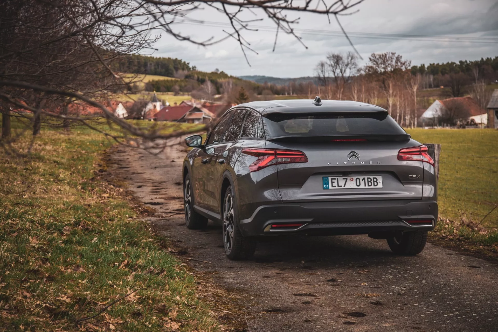 Vista trasera del hatchback Citroën C5-X 2022 en un camino de tierra con pueblo de fondo, mostrando luces traseras, parachoques trasero, doble escape, un emblema que indica que es un modelo Citroën, y un acabado gris metálico bajo iluminación nublada