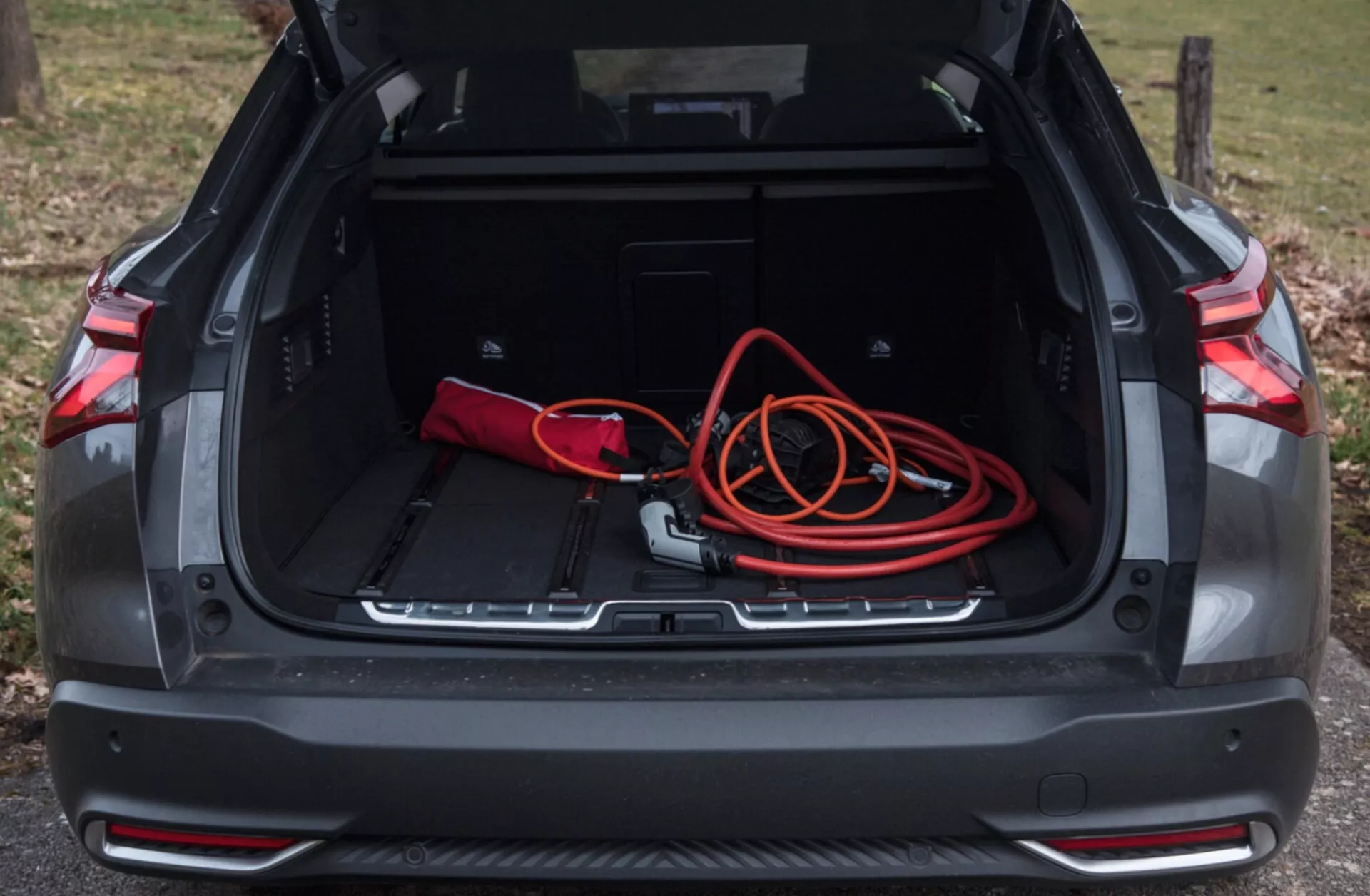 Vista del espacio del maletero trasero del Citroen C5-X 2022 con compartimento abierto mostrando área de carga espaciosa, bolsa roja y negra, cordón de extensión naranja, recesos de almacenamiento adicional, ribete protector, y sensores de estacionamiento, bajo iluminación natural al aire libre