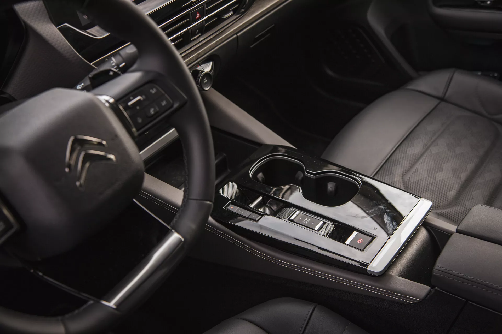 Vista interior del Citroën C5-X 2022 mostrando el volante multifunción con logo, tablero de alta tecnología con sistema de infoentretenimiento, palanca de cambios automática elegante, control climático, asientos tapizados en cuero con costuras de calidad y un toque de estilo de vida con gafas de sol en la superficie central brillante