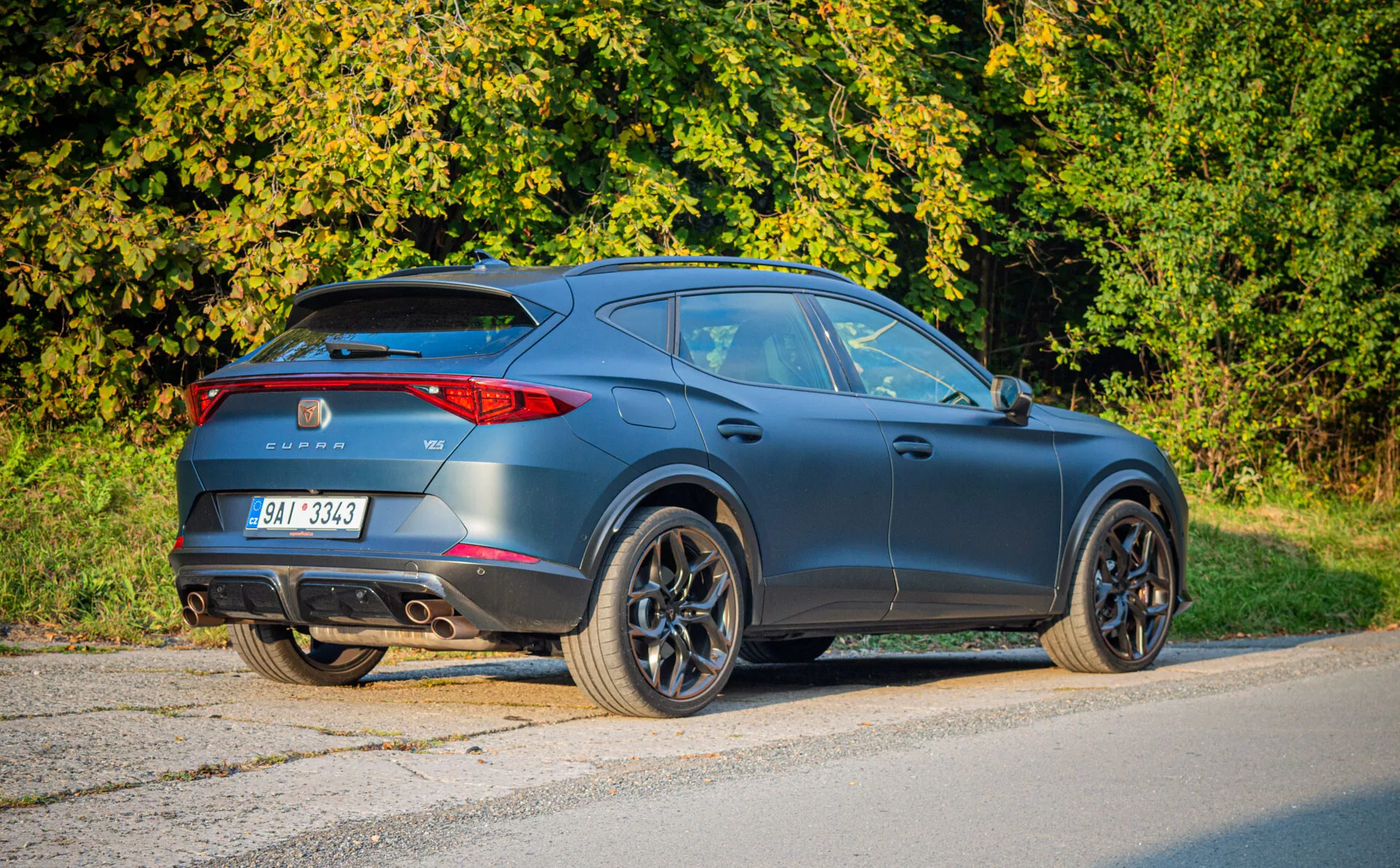 Moderno SUV crossover Cupra Formentor 2020 en gris oscuro mate negro estacionado en asfalto, mostrando vista trasera de tres cuartos resaltando diseño tipo coupé, aerodinámica deportiva, luces traseras LED, alerón trasero, escapes dobles y grandes ruedas de aleación en un entorno suburbano