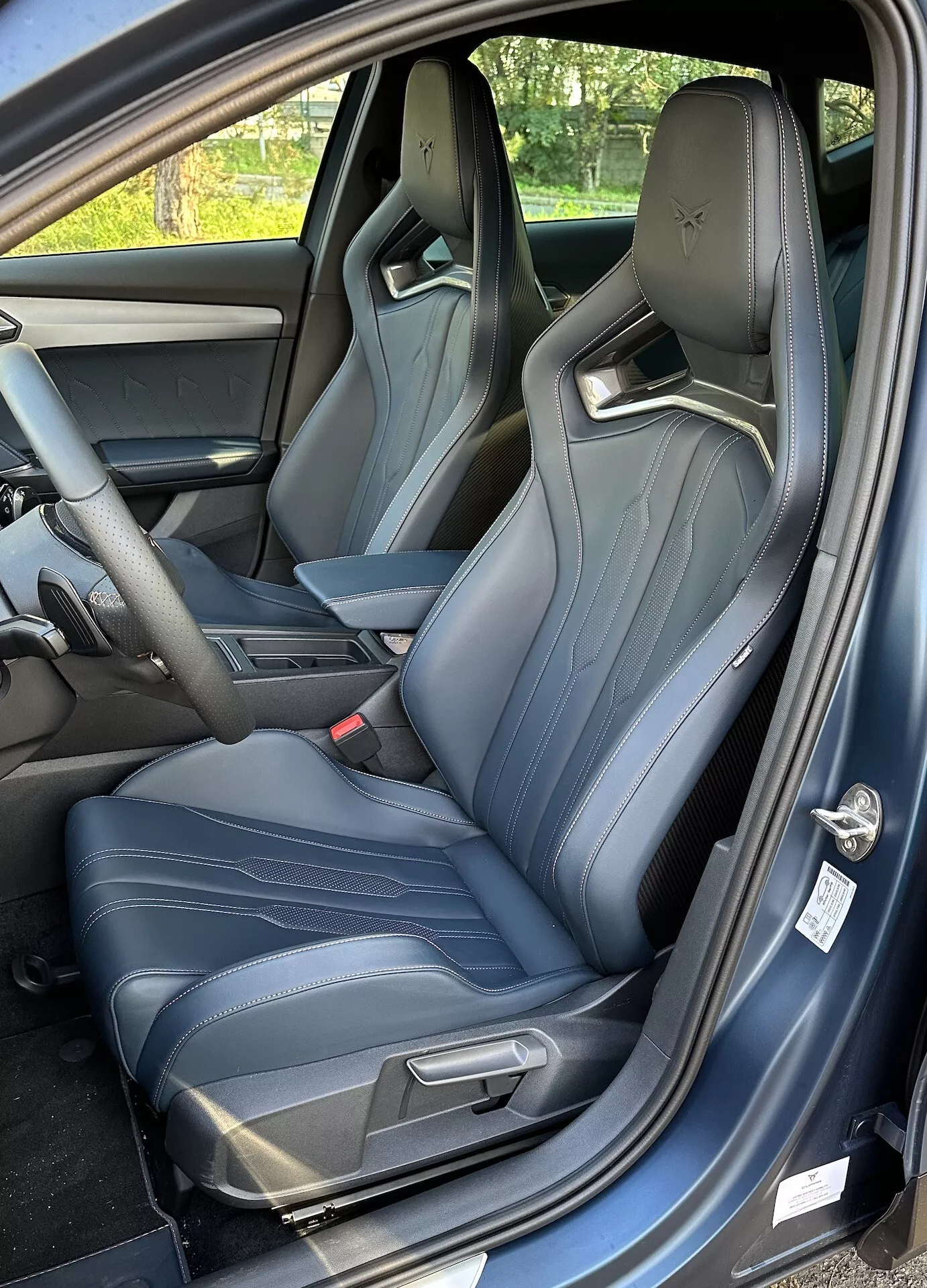 Vista interior de un Cupra Formentor 2020 mostrando el asiento del conductor desde una perspectiva de tres cuartos, con la puerta abierta, asientos tipo baquet deportivos con costuras en azul claro y un vistazo de la consola central y el tablero.