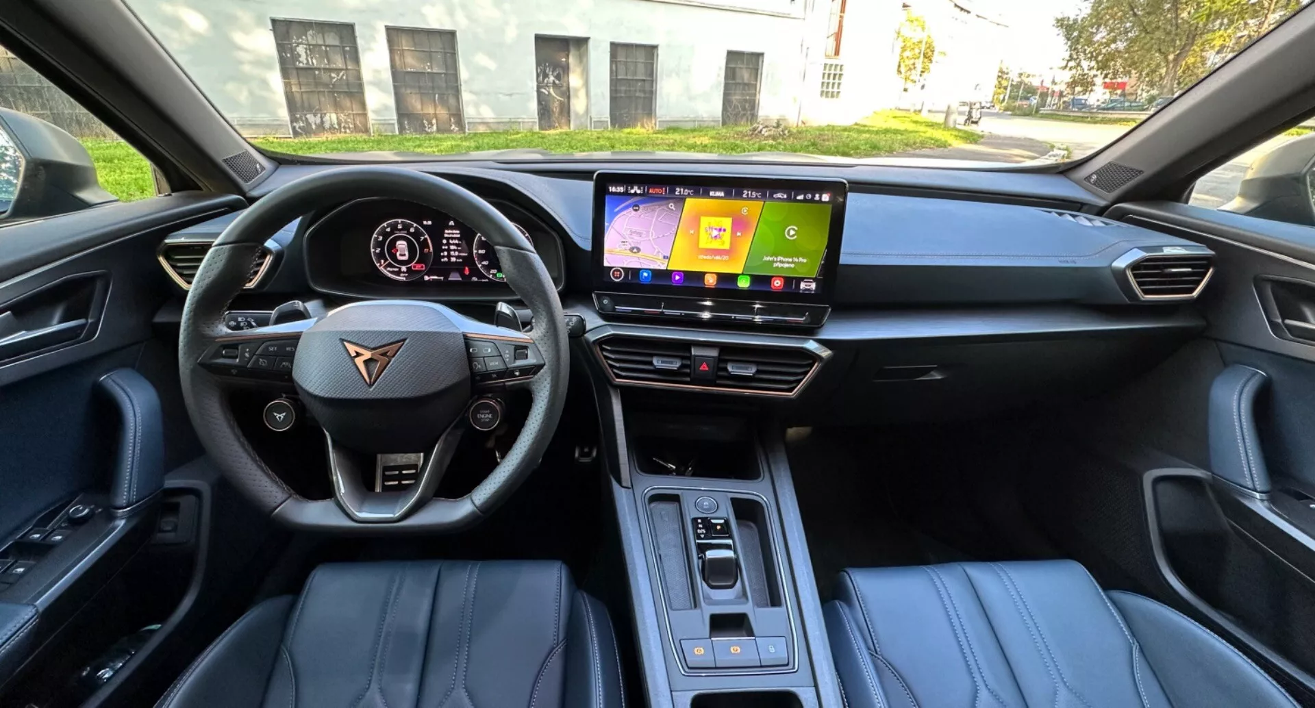 Vista interior desde la perspectiva del asiento del conductor de un CUPRA Formentor 2020 mostrando un volante de fondo plano con botones para multimedia y control de crucero, un tablero digital con visualización de velocidad y RPM, una pantalla de infoentretenimiento central, selector de cambios automático elegante, asientos tapizados en cuero con acento en dos tonos, e iluminación de la cabina diurna.