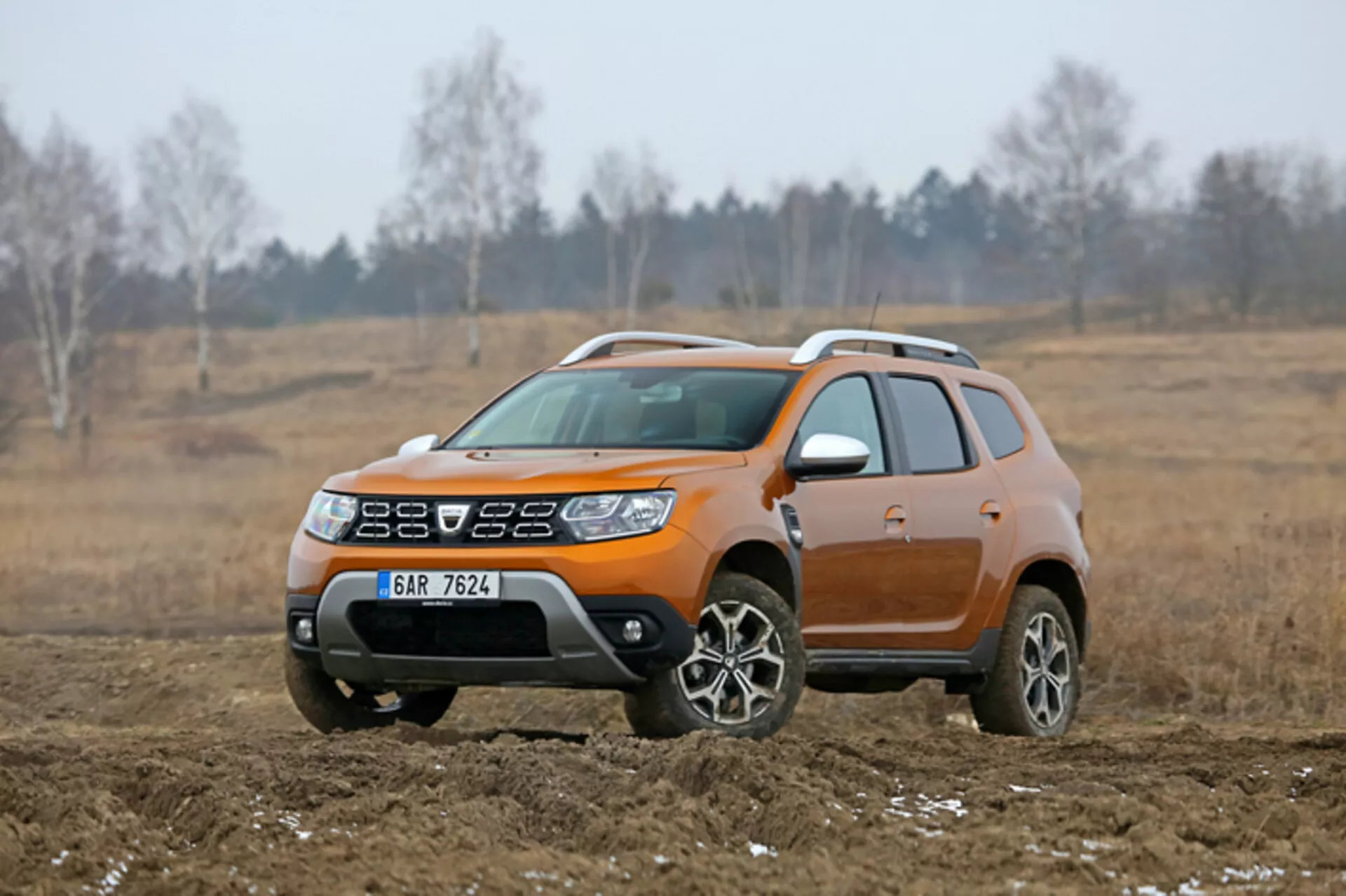 SUV compacto Dacia Duster 2013 exhibido en un entorno exterior todoterreno, destacado por su distintivo color naranja, altura elevada del viaje, ruedas de aleación y revestimiento de carrocería de plástico para uso todoterreno ligero, capturado desde un ángulo frontal de tres cuartos.