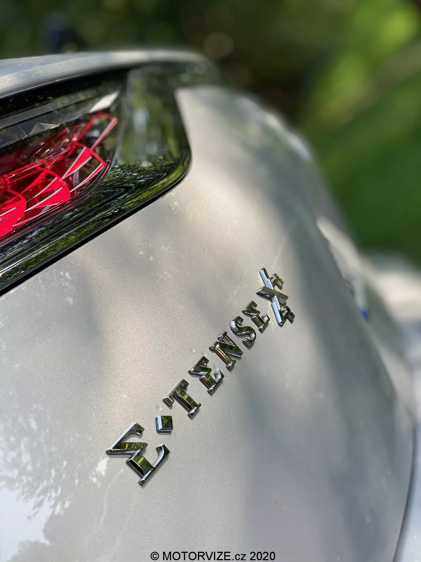 Vista en primer plano de la sección trasera del modelo DS Automobiles DS 7 Crossback 2018, centrándose en el emblema 'E-TENSE 4X4' que indica el sistema de tracción total y probablemente un tren de tracción híbrido o eléctrico. La poca profundidad de campo desenfoca el fondo mientras resalta el emblema, con la luz trasera cristalina roja visible arriba. La imagen captura el acabado metálico del cuerpo y el letreo cromado bajo la luz natural del sol, enfatizando la tecnología avanzada y el diseño lujoso del vehículo.