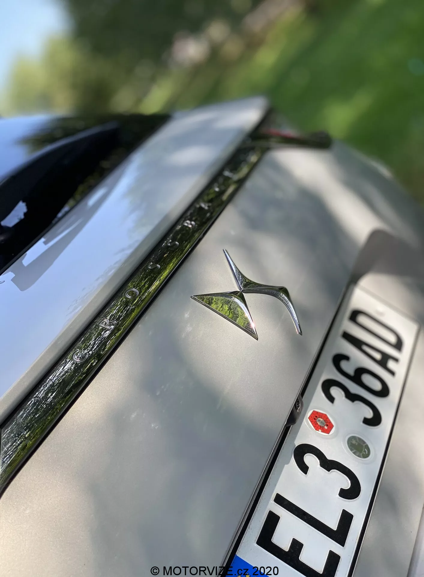 Foto en primer plano angulado de la parte trasera de un DS Automobiles DS 7 Crossback plateado de 2018. La imagen destaca el emblema metálico del coche centrado en la tapa del maletero, una matrícula europea con caracteres negros 'EL3 36AD' debajo del emblema y el borde superior del parachoques trasero. Parte del parabrisas trasero con el descongelador visible, junto con superficies reflectantes de alto brillo, también aparece. La profundidad de campo reducida enfatiza el emblema mientras desenfoca suavemente los bordes de la tapa del maletero y del parabrisas.
