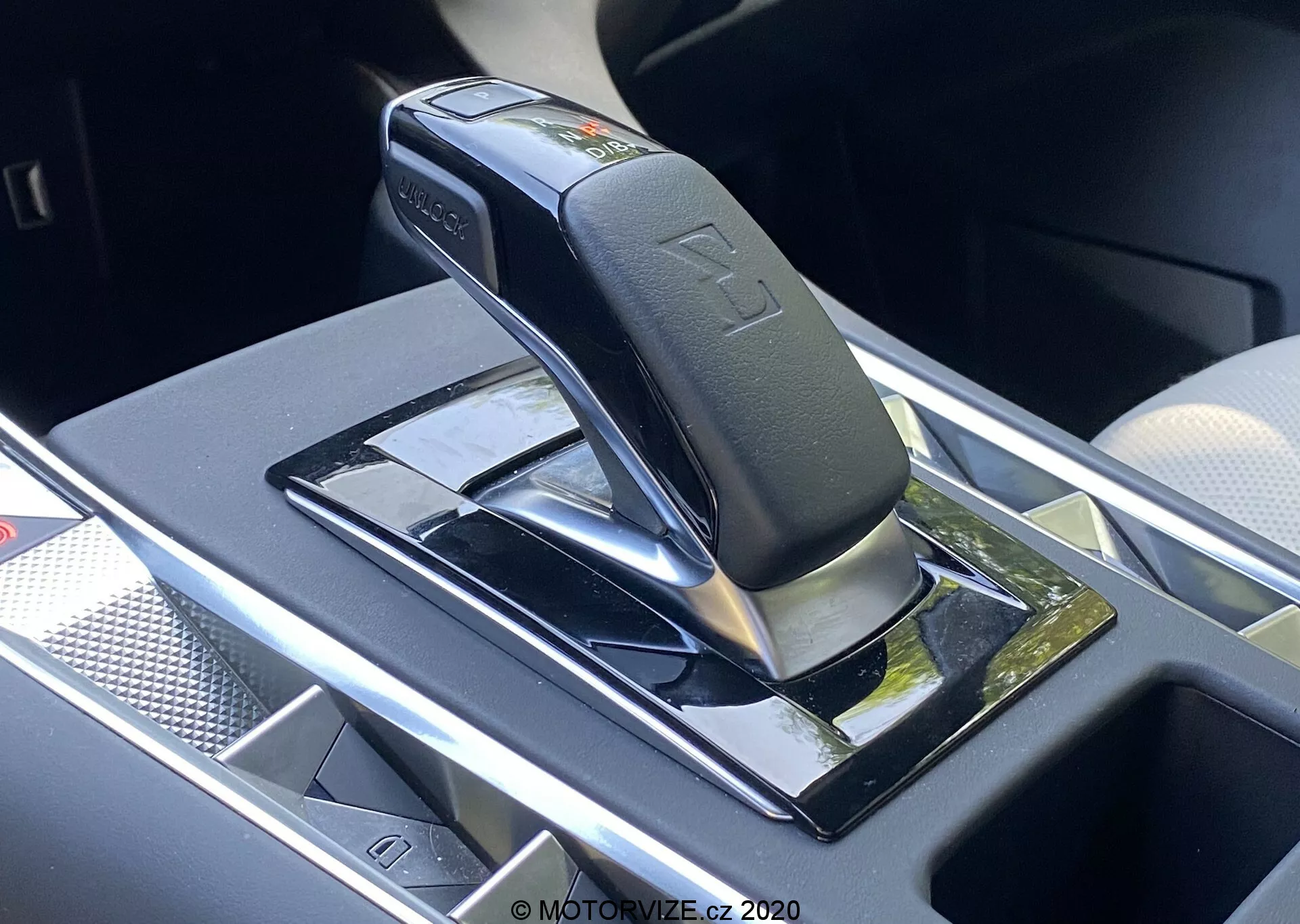 Vista en primer plano de la palanca de cambios automática del DS Automobiles DS-7 2018 (DS-7 Crossback), mostrando una palanca negra brillante con acentos plateados, probablemente en posición de estacionamiento o neutral, montada en un panel negro piano con superficies reflectantes. Las características incluyen símbolos en relieve de las posiciones de los cambios, un botón de seguridad en la palanca y un compartimento de almacenamiento cercano con una posible plataforma de carga inalámbrica. El área circundante tiene adornos cromados o plateados, lo que indica un interior de vehículo premium y moderno.