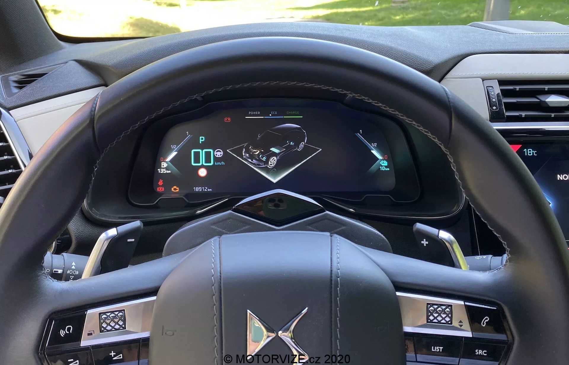 Vista de cerca del lado del conductor de un coche moderno desde la perspectiva del ocupante que muestra un volante multifuncional forrado en cuero con acentos metálicos y varios botones de control, un tablero de instrumentos totalmente digital que muestra velocidad, marcha y mapa de navegación, palancas de control para señales de giro y limpiaparabrisas en las columnas del volante, botones para el sistema de infoentretenimiento y climatización en el tablero, paletas de cambio semiautomáticas, un botón de arranque/parada, y materiales de alta calidad en el interior.