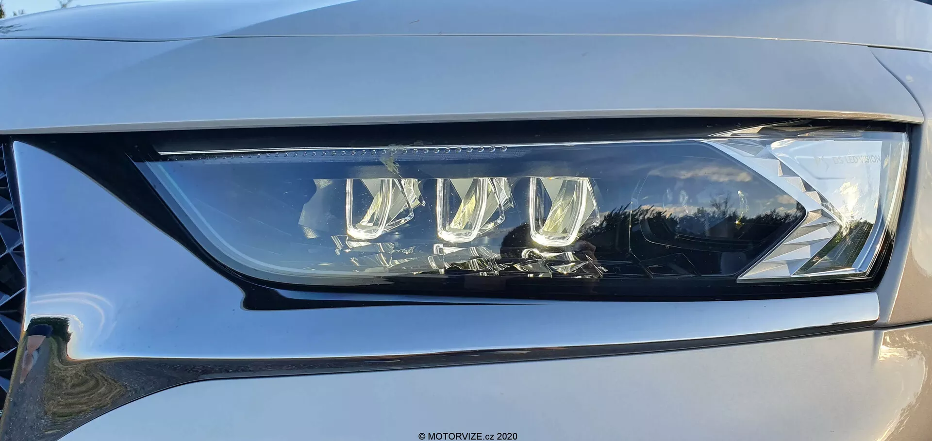 Primer plano del conjunto de faros izquierdo del DS Automobiles DS-7 Crossback desde un ángulo leve, que muestra un diseño LED moderno con múltiples lentes de proyector y una distintiva luz diurna LED, encapsulada en plástico transparente de alta calidad. El faro ejemplifica tecnología avanzada de iluminación de vehículos, capaz de proporcionar funciones de luz de cruce y de carretera, con posibles características adaptativas para mejorar la visibilidad. También se puede ver un reflejo sutil en la superficie brillante y un toque del color gris claro a medio del coche.