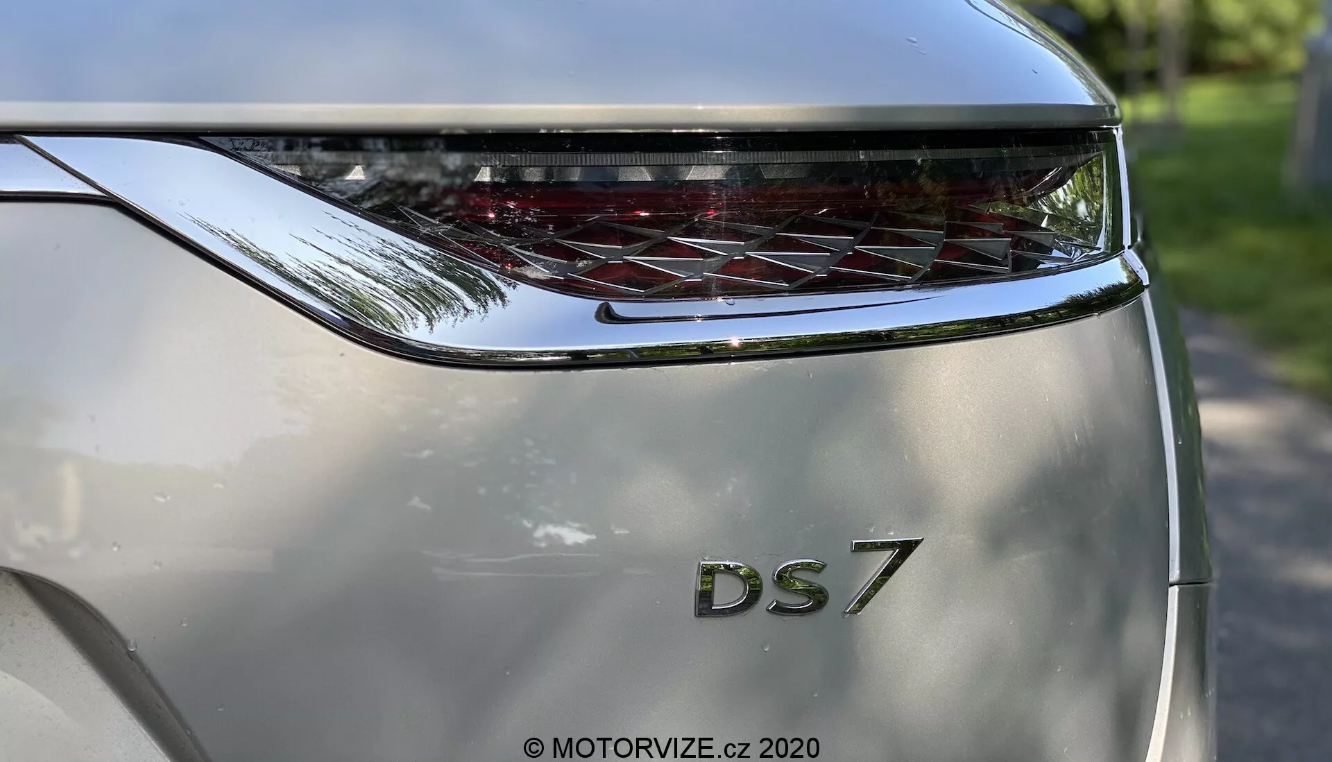 Vista de cerca de la sección trasera de un DS Automobiles DS 7 Crossback 2018 capturada desde una perspectiva de ángulo bajo. El enfoque está en el portón trasero, que presenta destacadamente las letras en negrita 'DS' sobre la designación del modelo '7'. Sobre el nombre del modelo se encuentra una luz trasera curva y extendida horizontalmente con elementos LED distintivos. El color de la carrocería es plateado metálico y el diseño esculpido sugiere una apariencia moderna y lujosa. La fotografía enfatiza la marca y el diseño de las luces traseras, con un entorno exterior brillante reflejado sutilmente en la superficie del coche.