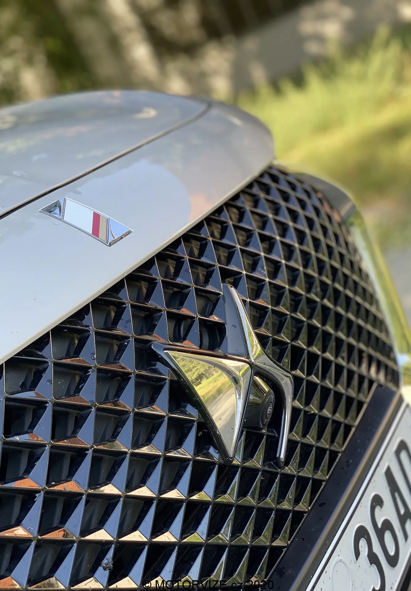 Primer plano de la sección delantera izquierda del DS Automobiles DS 7 Crossback 2018. La imagen se centra en la lujosa parrilla delantera con un intrincado patrón de diamantes y superficies reflectantes, centrada con un logo estilizado y prominente. Sobre la parrilla, el capó muestra un emblema más pequeño que indica un modelo de alta gama. El fondo está desenfocado, resaltando el diseño de la parrilla y los elementos de la marca.