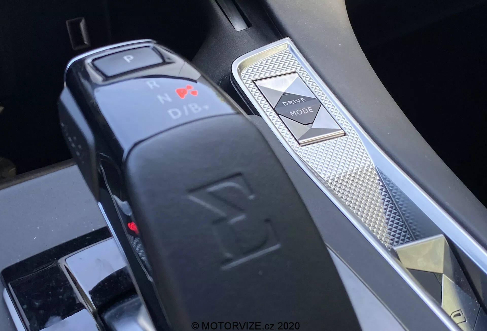 Vista de cerca de la consola central en un DS 7 Crossback 2018 que muestra el selector de marchas y el interruptor de modos de conducción desde un ángulo oblicuo. El selector de marchas electrónico, con un diseño en forma de L, resalta la posición de Parque con una luz LED roja y una función de botón. A la derecha, un interruptor de modos de conducción con patrón de diamante y un borde plateado indican el acabado premium del interior y los sistemas avanzados de control del vehículo.