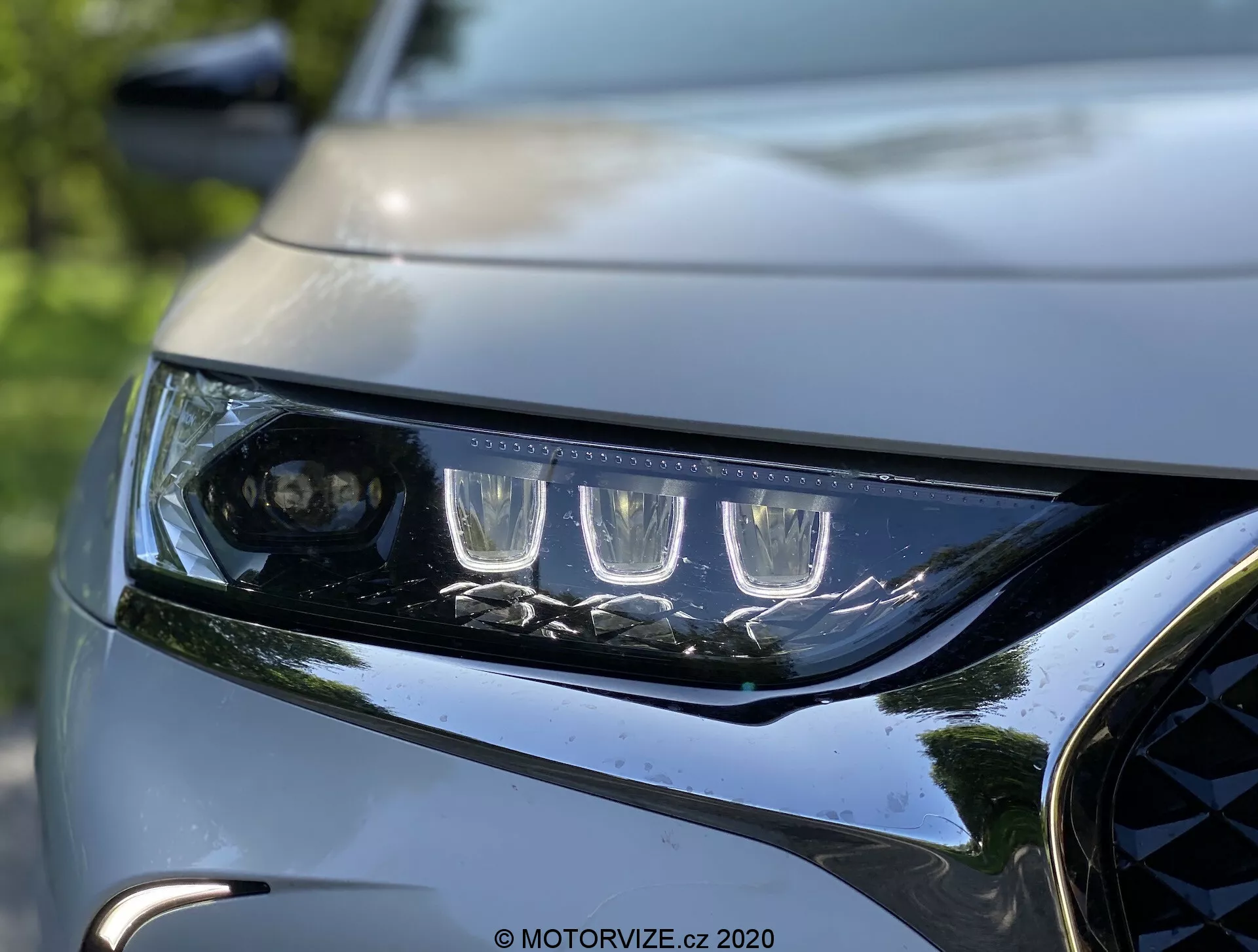 Vista de cerca del faro izquierdo y la parrilla frontal del DS Automobiles DS 7 Crossback 2018. El faro presenta luces diurnas LED distintivas y una lente proyectora para el haz principal, enmarcado por múltiples elementos LED. La carcasa del faro, elegante y angular, muestra una aerodinámica moderna. Una porción de la parrilla oscura y brillante con acentos metálicos y parte del capó contorneado resaltan el diseño dinámico y premium del vehículo. El fondo está desenfocado, destacando la nitidez y el detalle intrincado de la iluminación y la fascia frontal del coche.