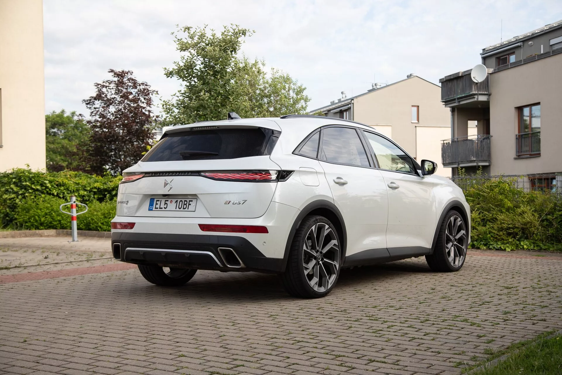 Vista trasera tres cuartos de un DS Automobiles DS 7 2022 en blanco, mostrando el perfil lateral y trasero con distintivas luces traseras horizontales estrechas, insignias, spoiler alto, antena aleta de tiburón, sensores de aparcamiento, sistema de doble escape y grandes ruedas de aleación de múltiples radios en una calle residencial.