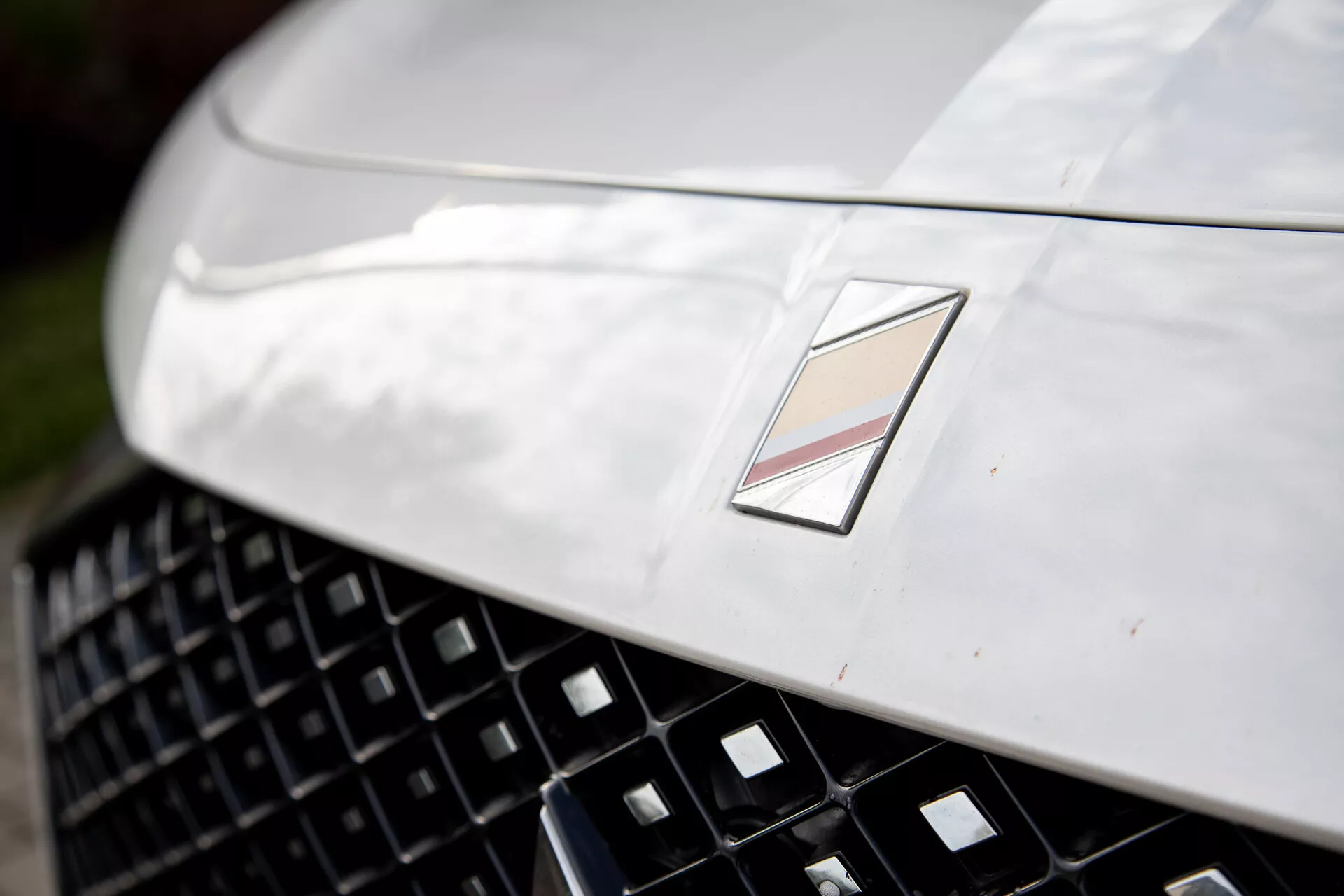 Vista cercana de la sección frontal de un coche de lujo mostrando un capó metálico con diseño aerodinámico, una rejilla detallada para una mejor entrada de aire, y un emblema multifacético que indica el fabricante, bajo reflejo de luz natural y profundidad de campo somera